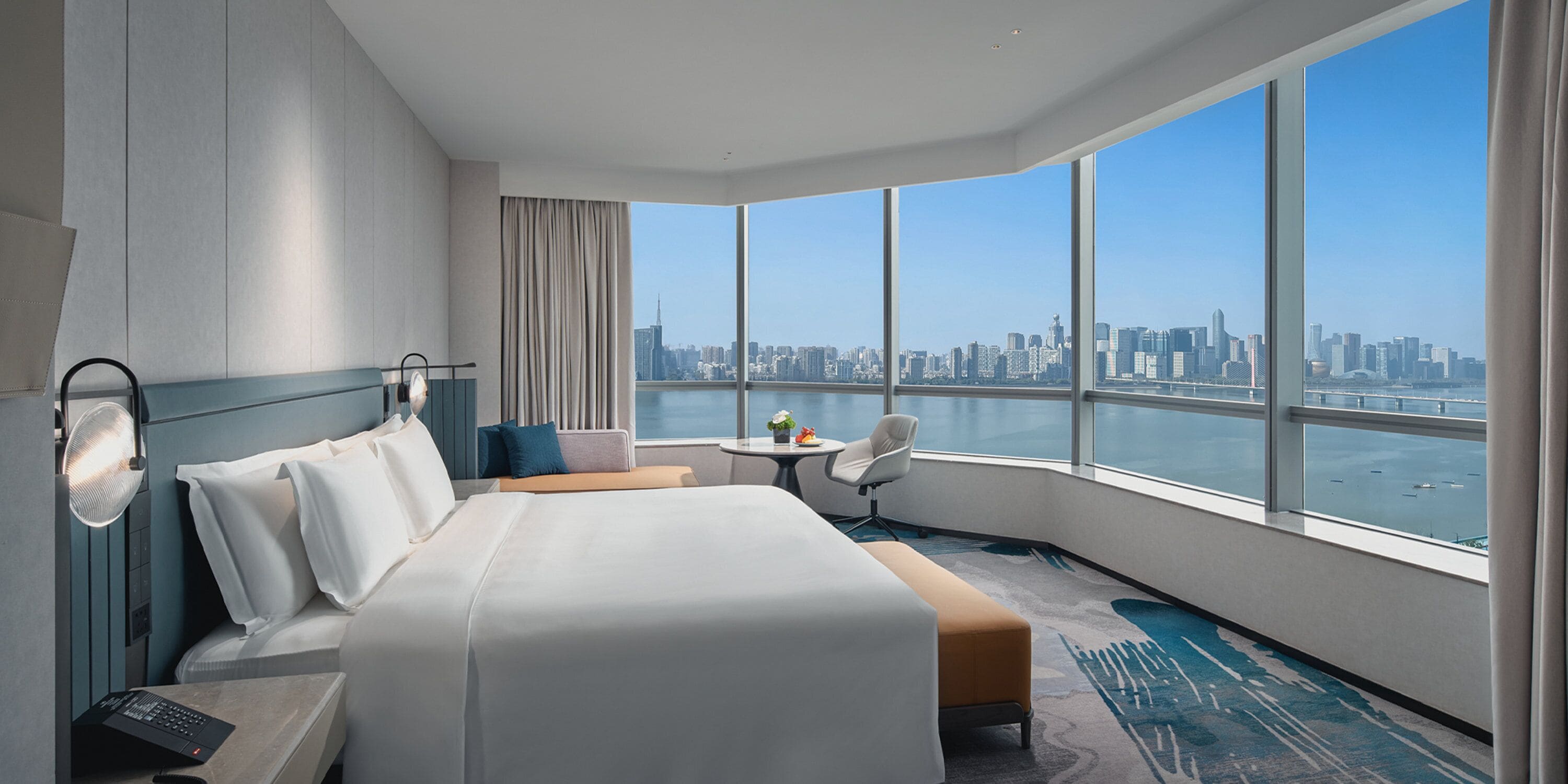 A hotel room with a large bed, a small table, a chair, and a view of a city skyline through large windows.