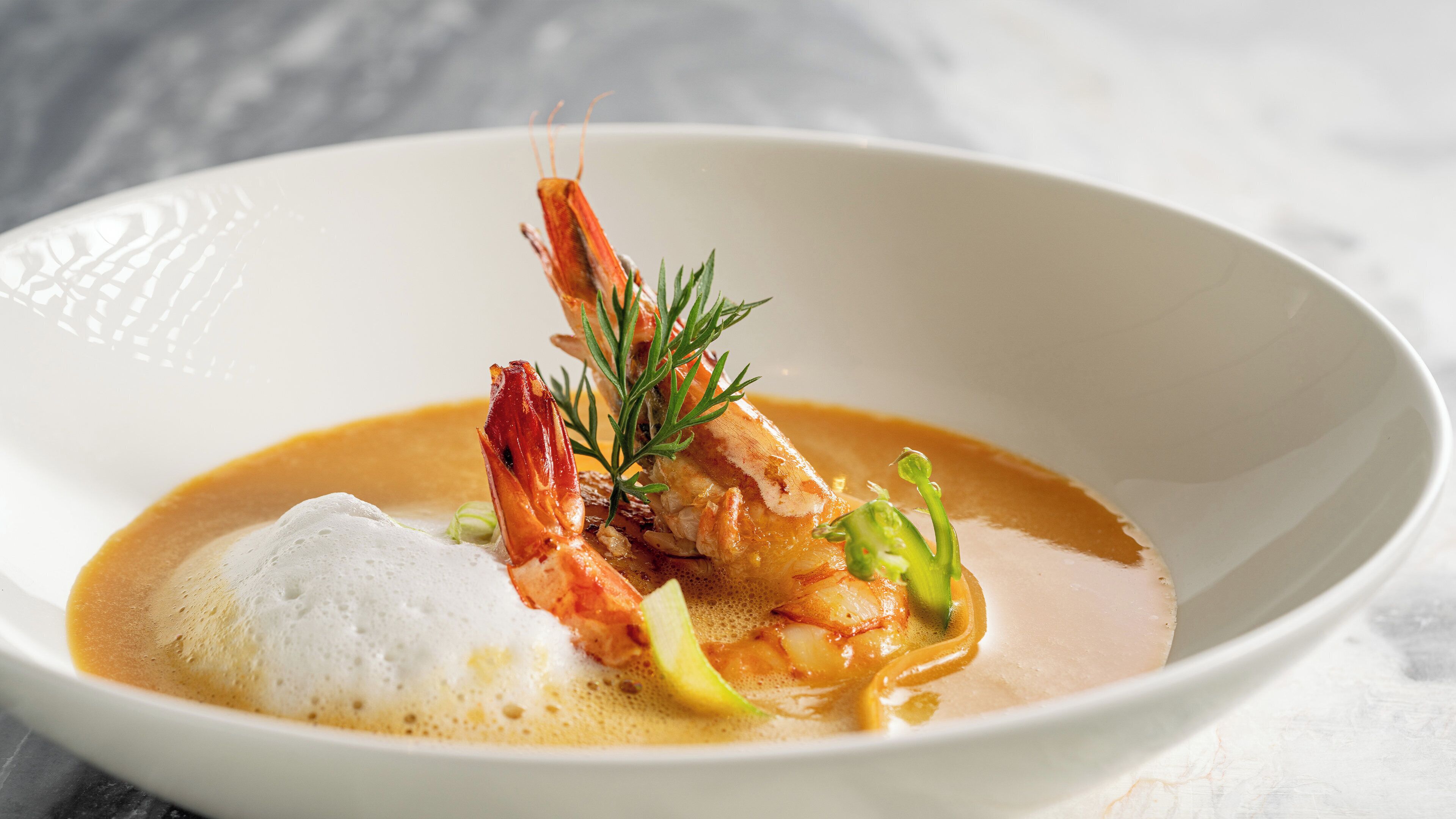 A white bowl containing a seafood dish with shrimp, a dollop of foam, and a garnish of green herbs.