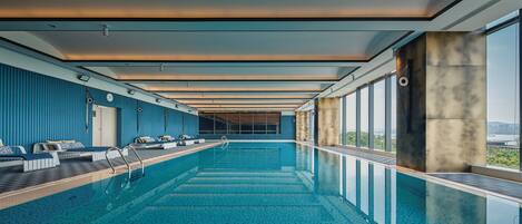 Una piscina interna moderna con vista sul cielo, un soffitto illuminato dal sole e un’area relax a bordo vasca con posti a sedere e una sveglia.