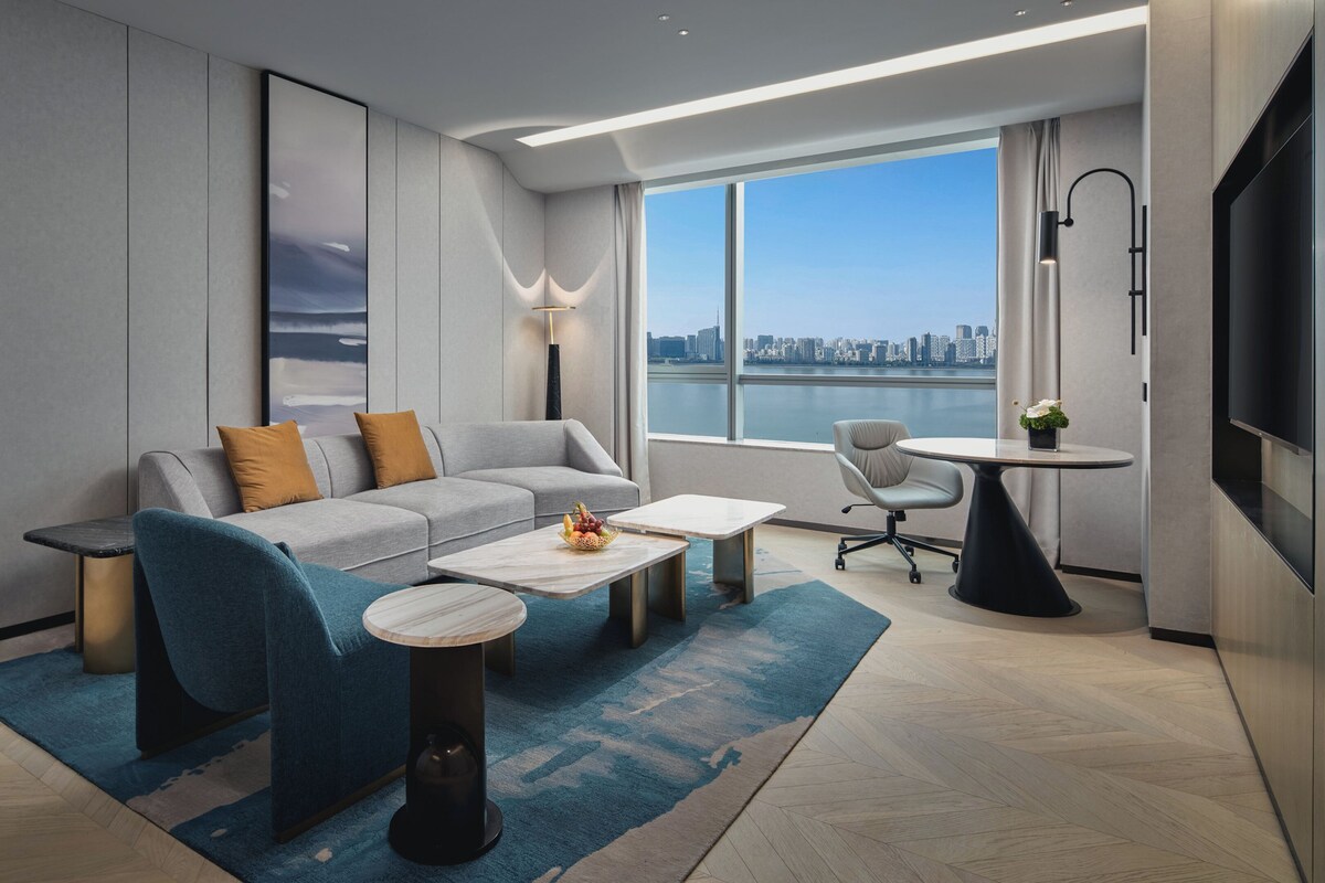 Quarto de hotel moderno com sofá, mesa de centro e vista para o horizonte da cidade.