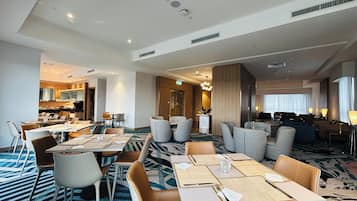 A hotel dining area with multiple tables and chairs, a kitchen in the background, and a living area with sofas and a TV.
