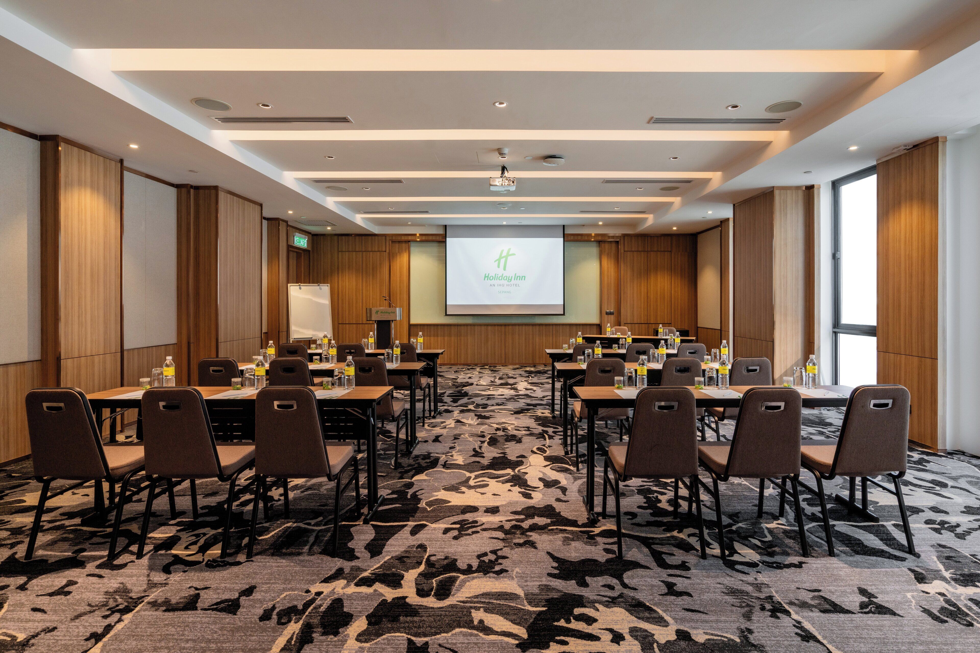 Un salón de conferencias con una pantalla grande mostrando la marca "Holiday Inn", varias mesas con sillas y una alfombra estampada.
