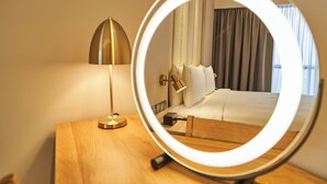 A hotel room with a wooden bedside table, a lamp, a round mirror with a light, and a bed with white linens. - Holiday Inn Sepang Airport by IHG (Sepang)