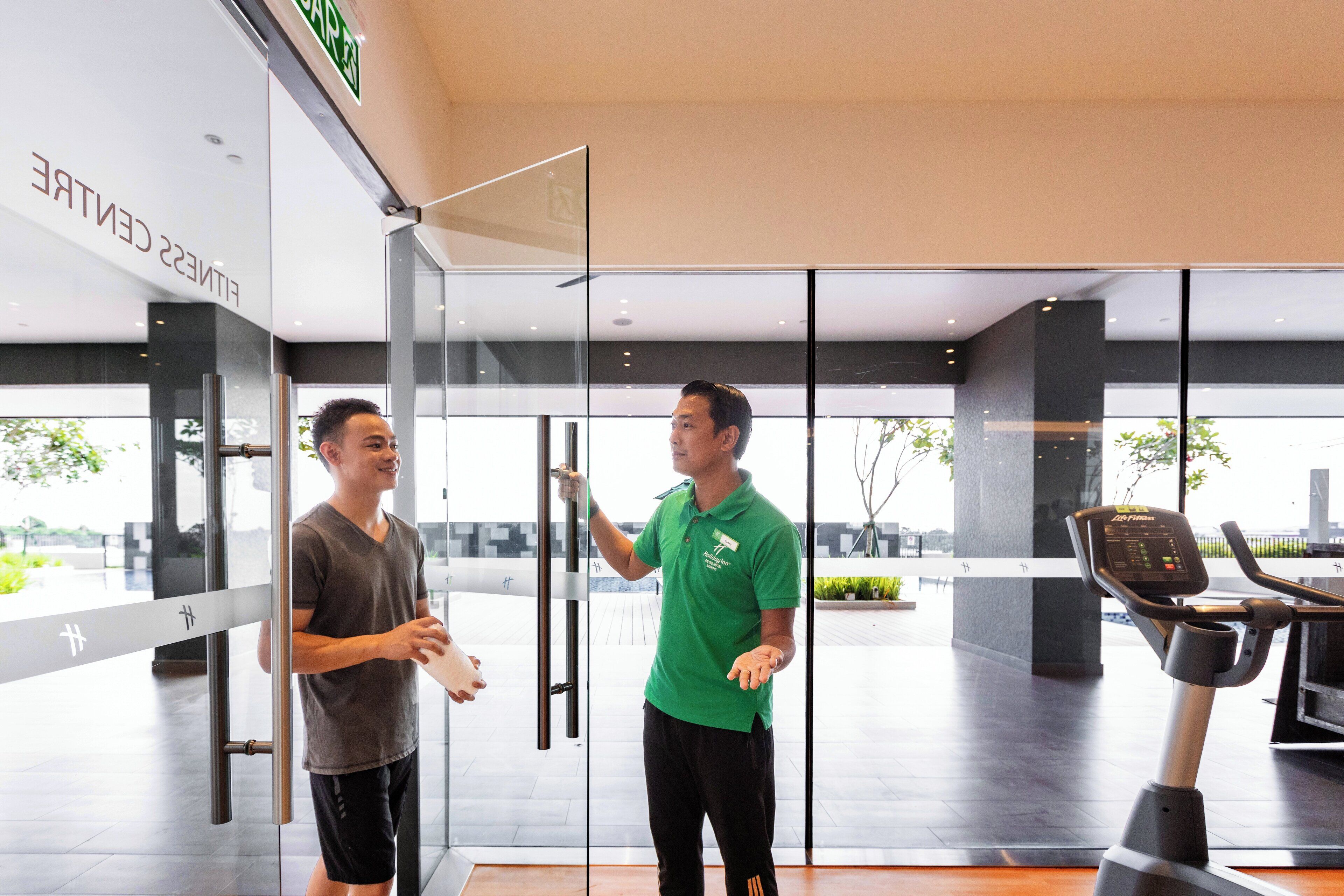 Dos personas en un gimnasio moderno con puertas de vidrio, una cinta de correr y vistas al exterior.
