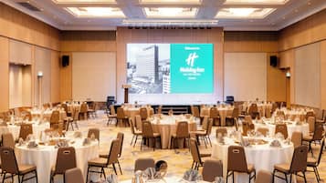 Ein großer Konferenzraum mit mehreren runden Tischen, gedeckt für eine formelle Veranstaltung, eine große Leinwand mit einer Willkommensbotschaft und einem modernen Deckenstil.