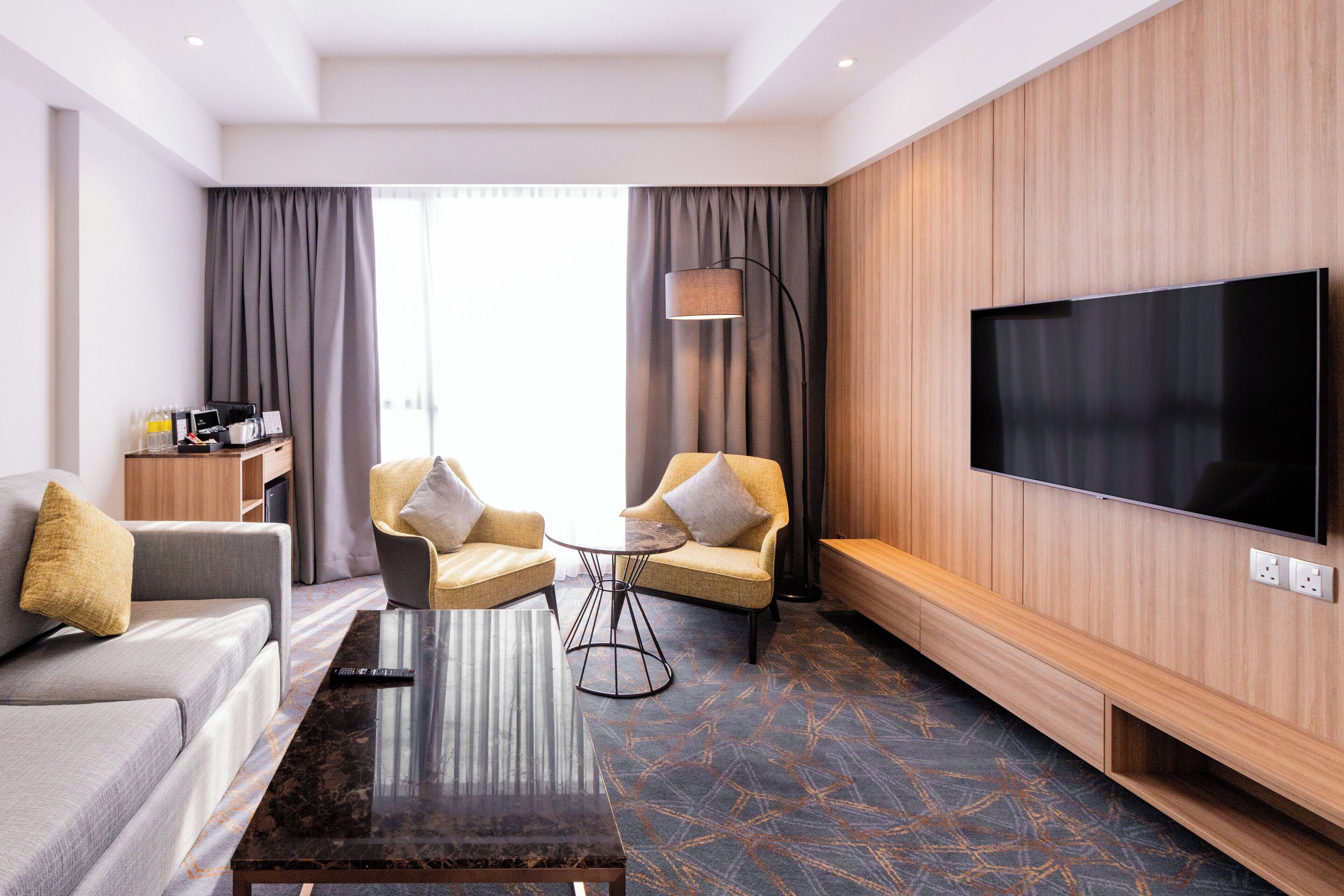 A modern hotel room with a sofa, two armchairs, a coffee table, a flat-screen TV, and a wooden accent wall.