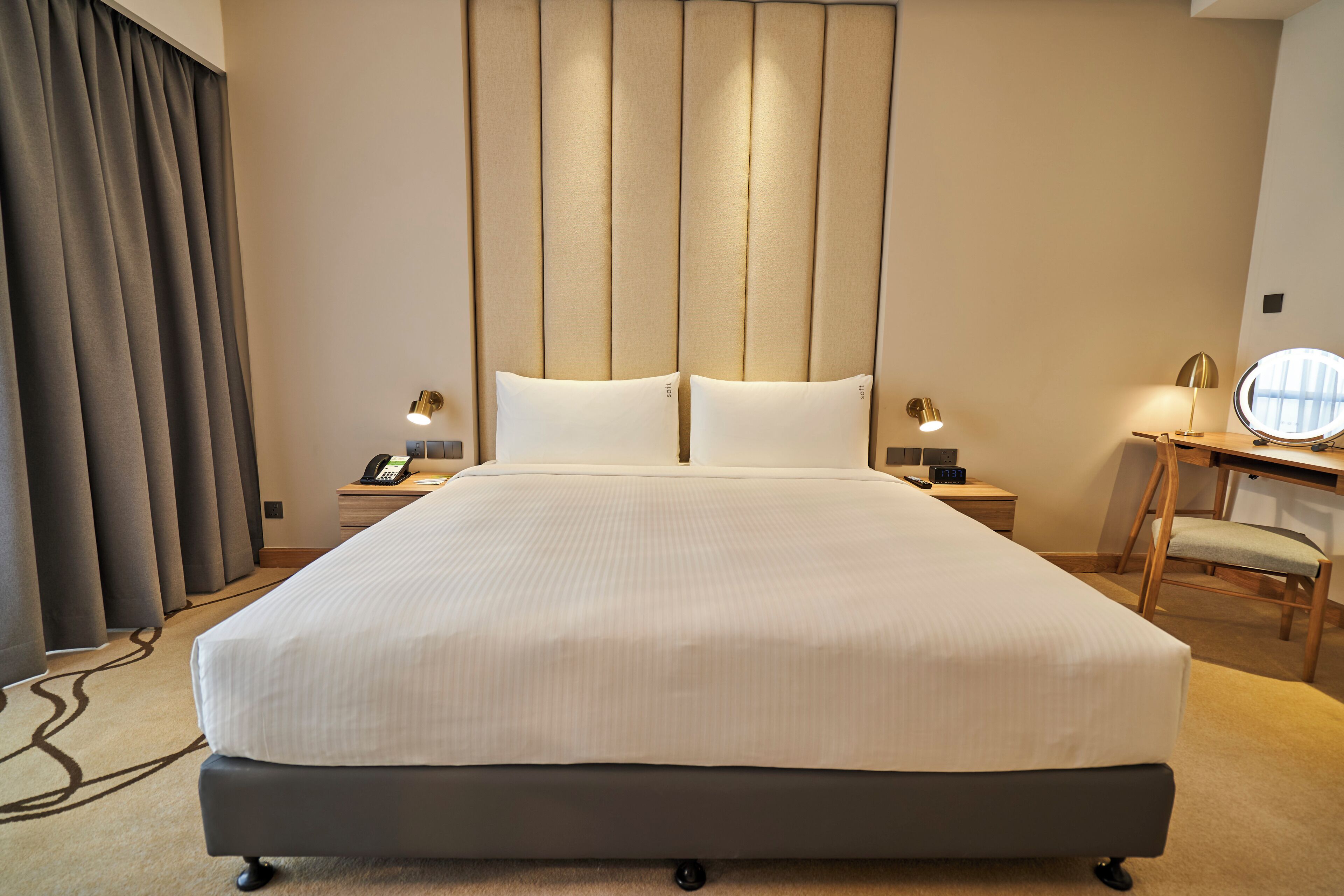 Quarto de hotel com uma cama grande, mesas de cabeceira com luminárias, uma escrivaninha com cadeira e uma janela ampla com cortinas.