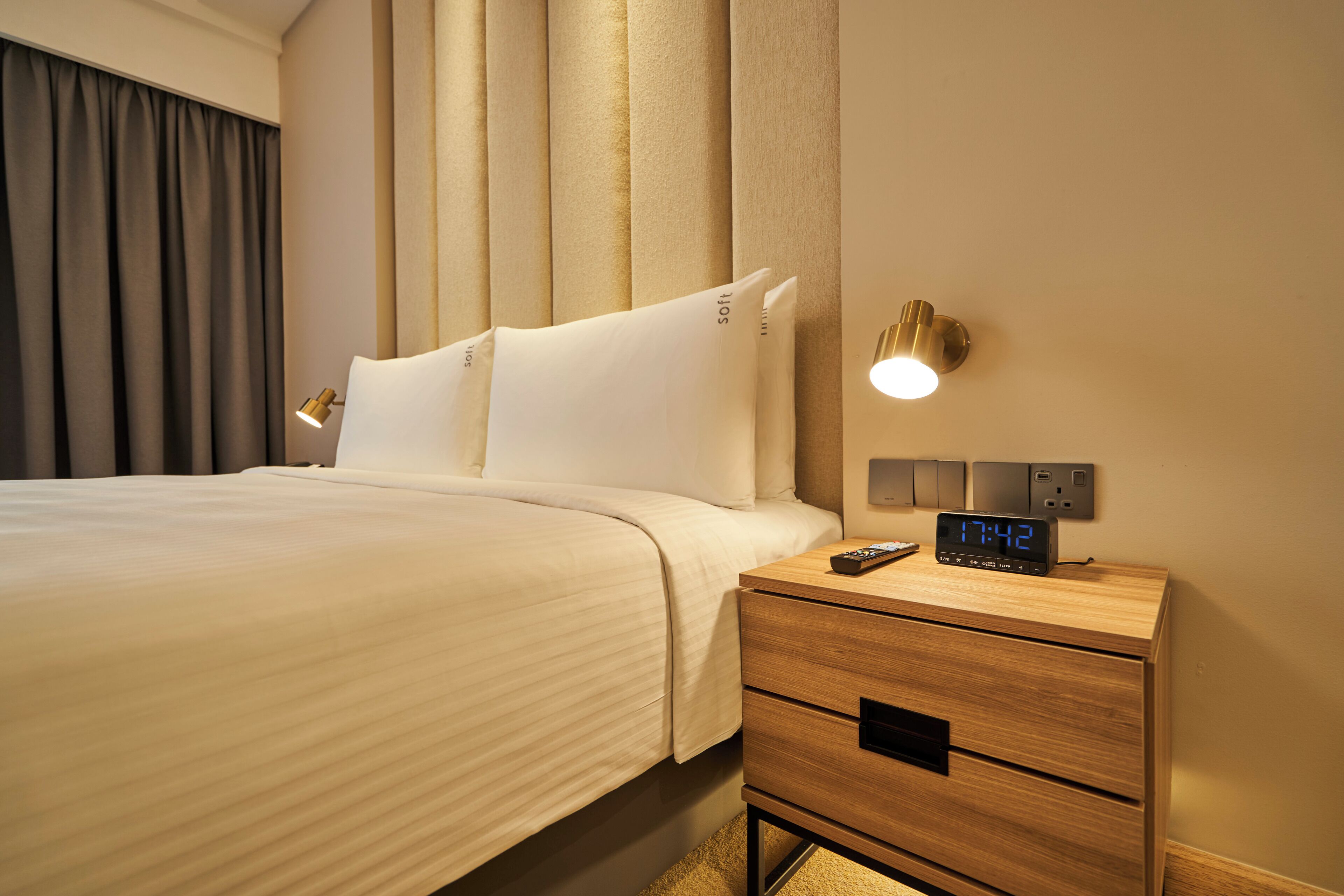 A hotel room with a bed, a bedside table with a digital clock, and a wall-mounted light.