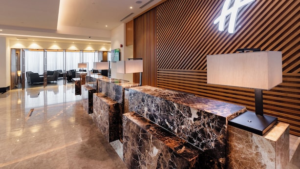 A modern hotel lobby with a marble reception desk, wooden paneling, and a large illuminated 'H' sign. - Holiday Inn Sepang Airport by IHG (Sepang)