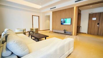 A modern hotel room with a large sofa, a small table, a TV mounted on the wall, and a wooden door.