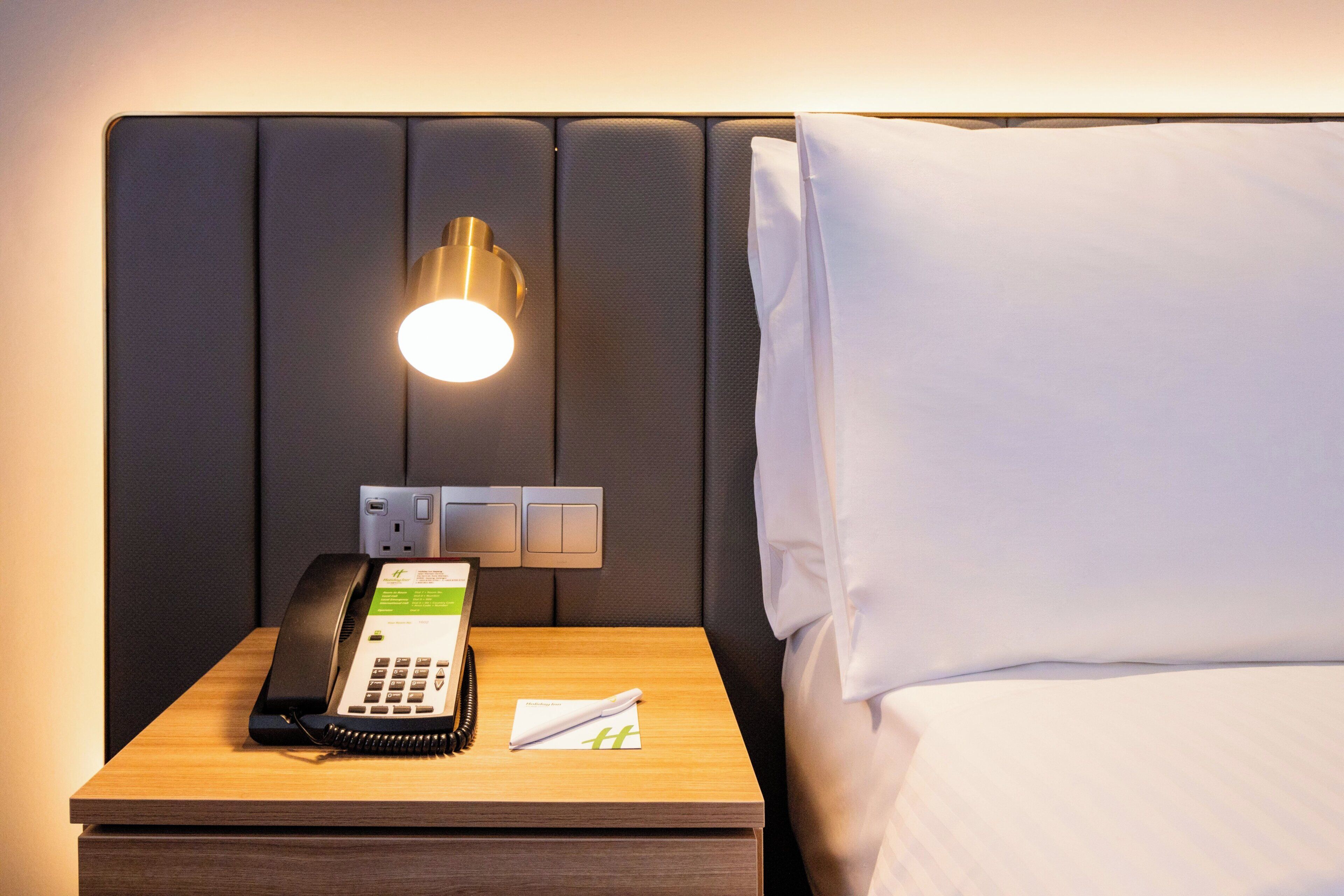 A hotel room with a bedside table, a telephone, a wall-mounted light, and a bed with white bedding.