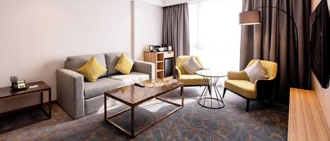 A modern hotel room with a grey sofa, yellow armchairs, a coffee table, a TV, and a bookshelf.