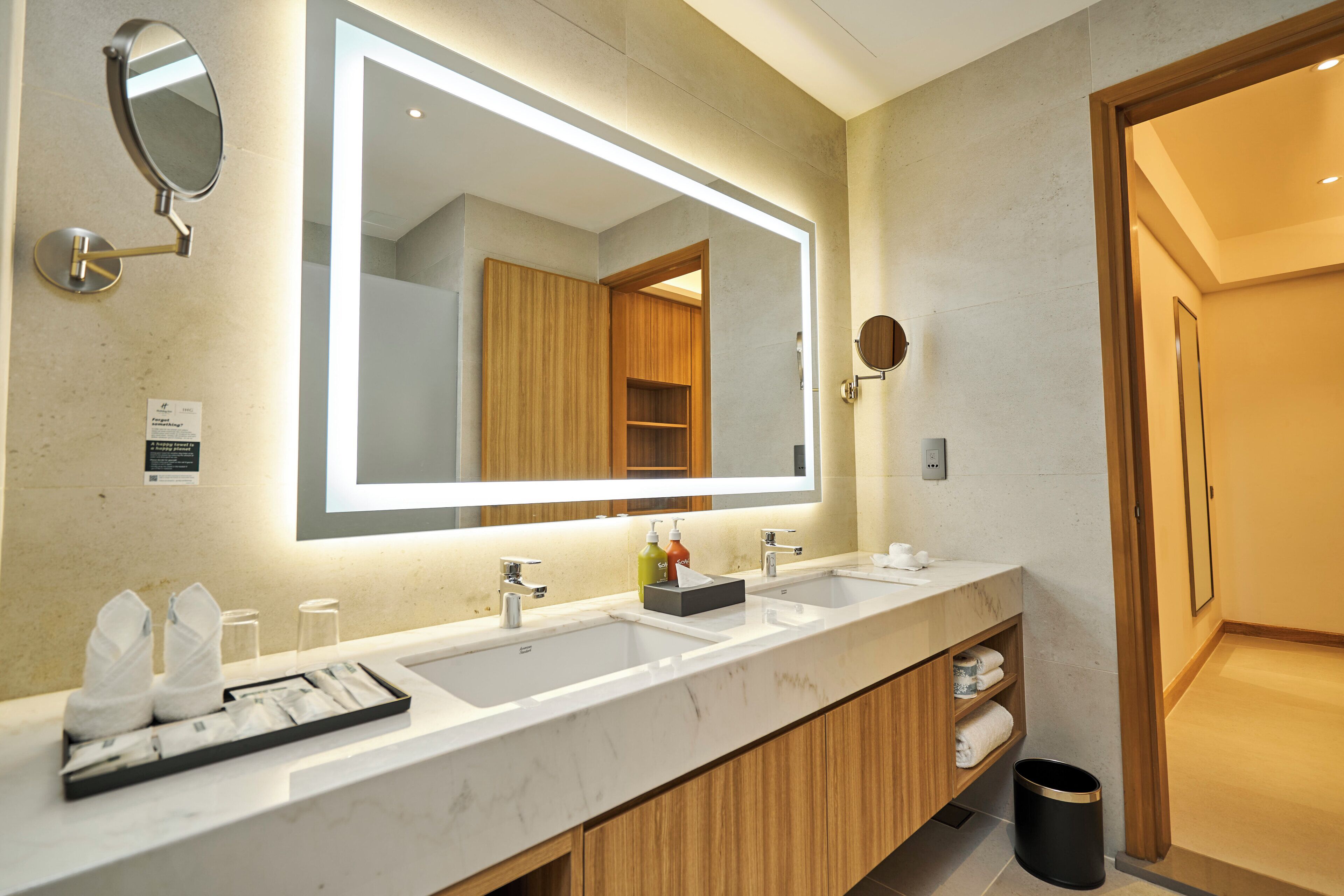 A modern bathroom with a large mirror, two sinks, wooden cabinets, and a trash can.