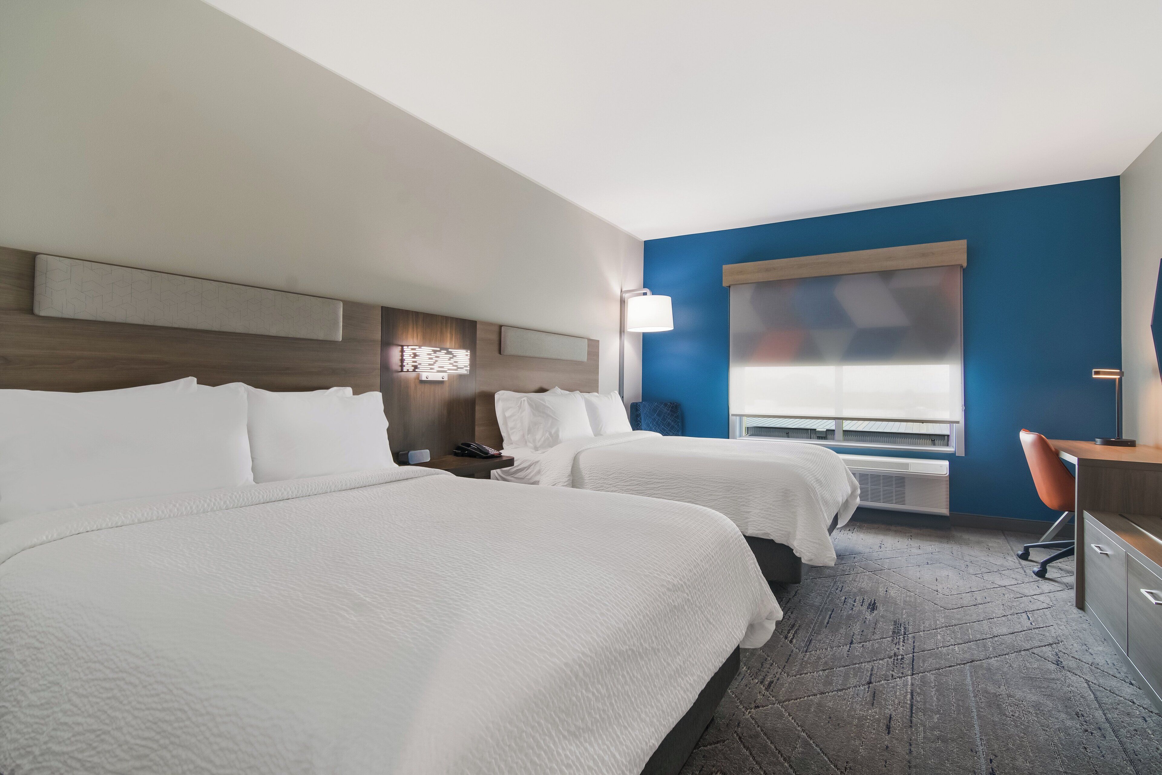 A hotel room with two beds, a desk, a chair, a nightstand, and a window with blinds.