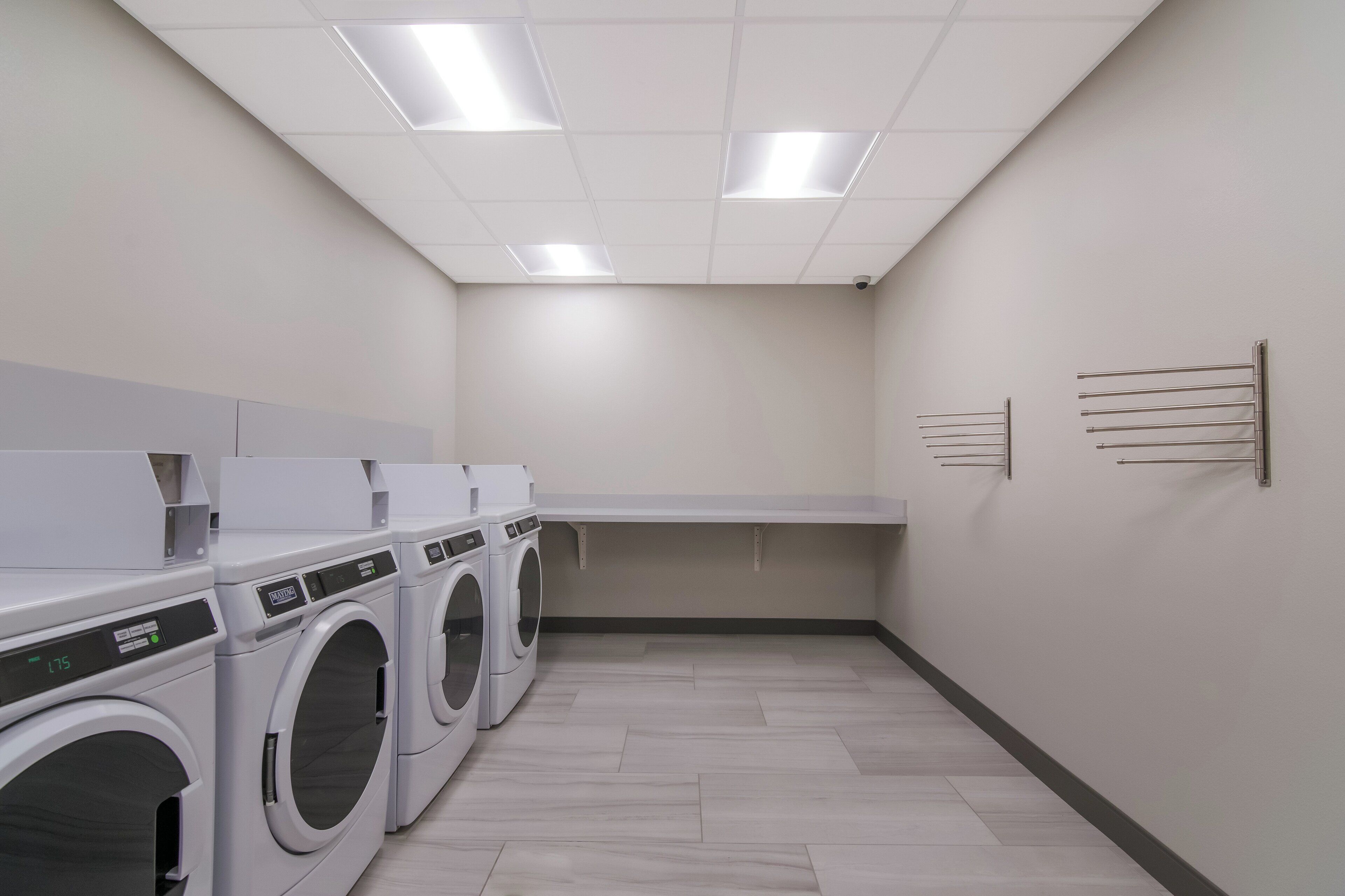 A laundry room with multiple washing machines and dryers, a tiled floor, and a plain wall with a shelf.