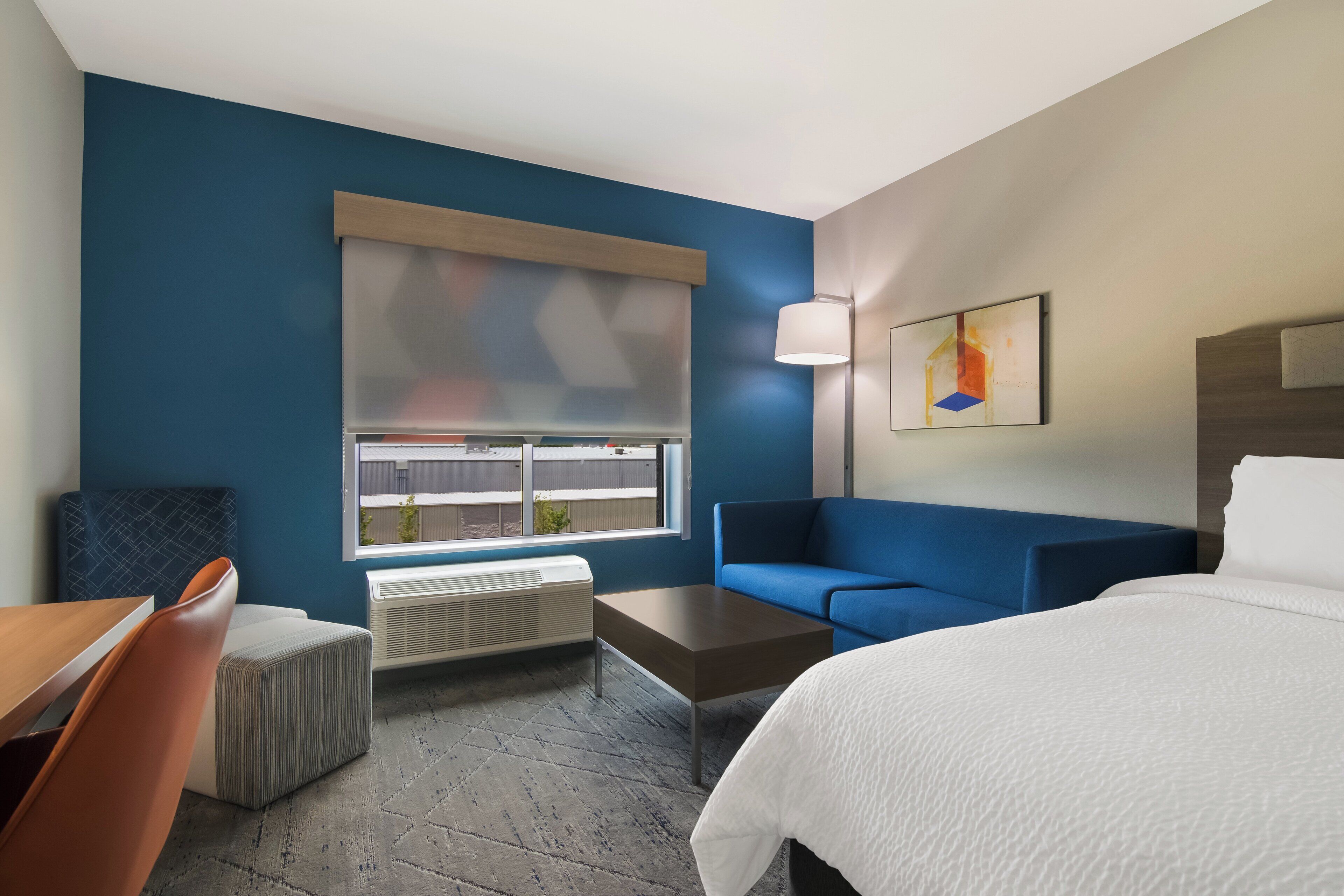 A hotel room with a bed, a blue sofa, a wooden table, and a window with blinds.