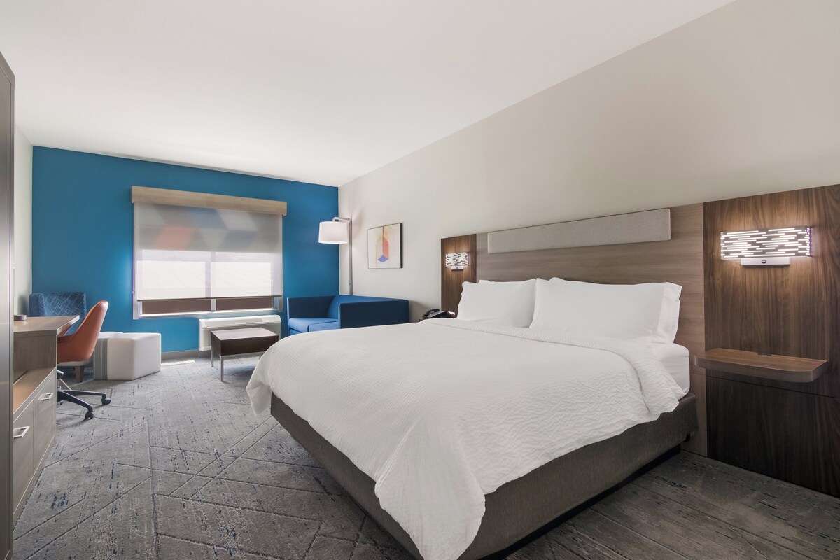 A modern hotel room with a large bed, a desk with a chair, a blue accent wall, and a window.