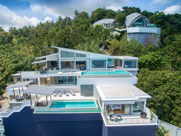 A multi-story modern house with a large swimming pool, multiple balconies, and lush surrounding vegetation.