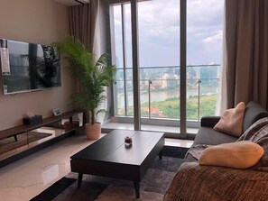 A modern living room with a large window, a flat-screen TV, a coffee table, a sofa, and a potted plant. - Hoasun Emperor Apartment - Empire City (Ho Chi Minh City)