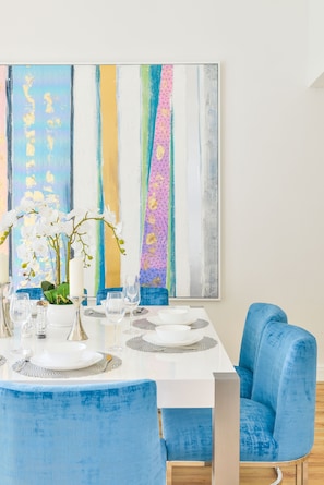 A dining area with a white table set for four, blue upholstered chairs, and a colorful abstract painting on the wall. - Al Bateen - Ultra Luxury JBR - Private Beach and Pool (Dubai)
