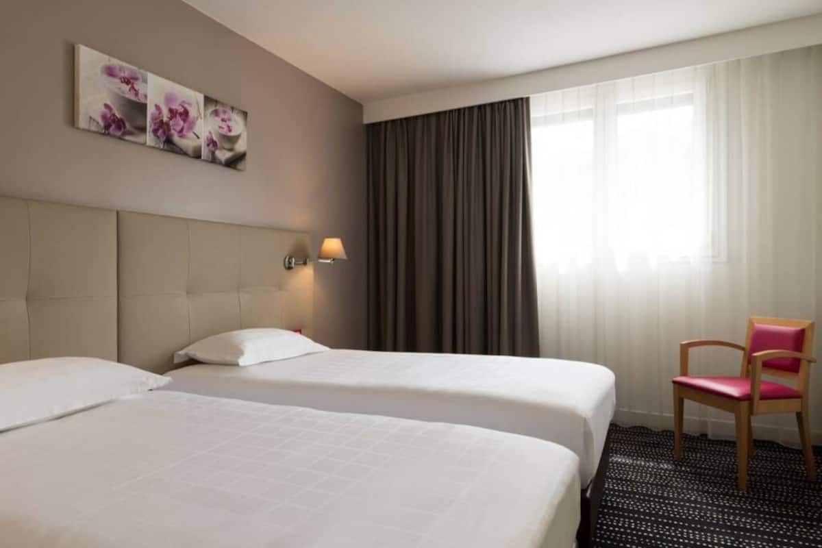 A hotel room with two beds, a chair, a painting, and a window with curtains.