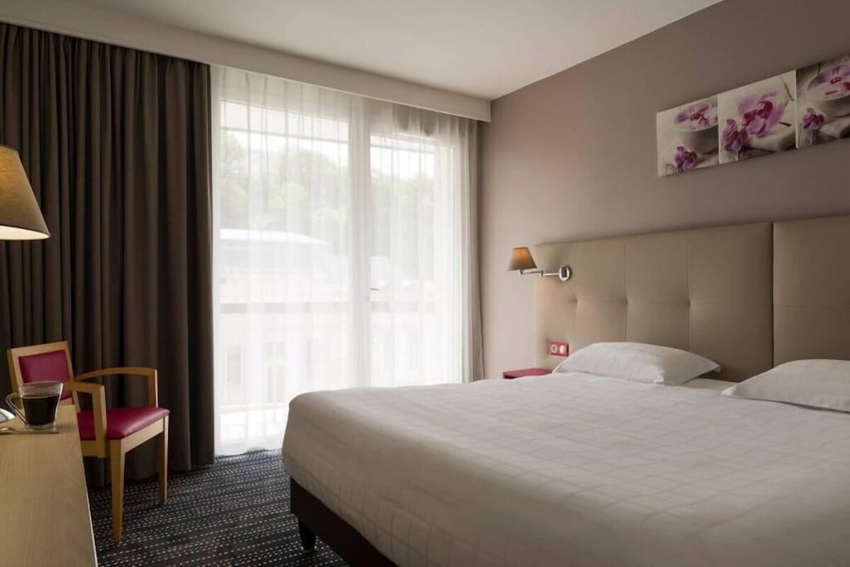 Una habitación de hotel con una cama, una silla, un escritorio con lámpara y una ventana con cortinas.