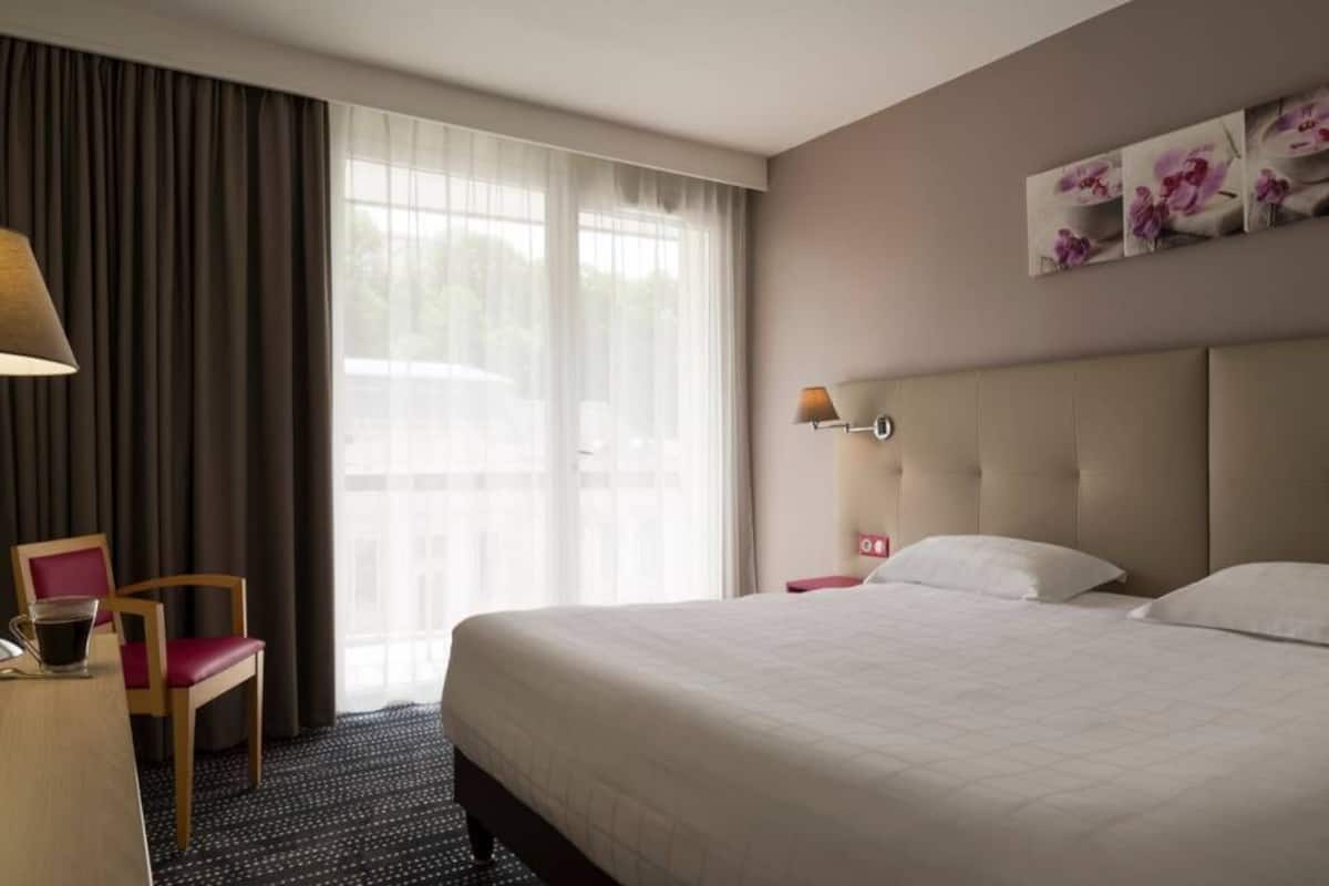 A hotel room with a large bed, a desk with a chair, a window with curtains, and a wall-mounted lamp.