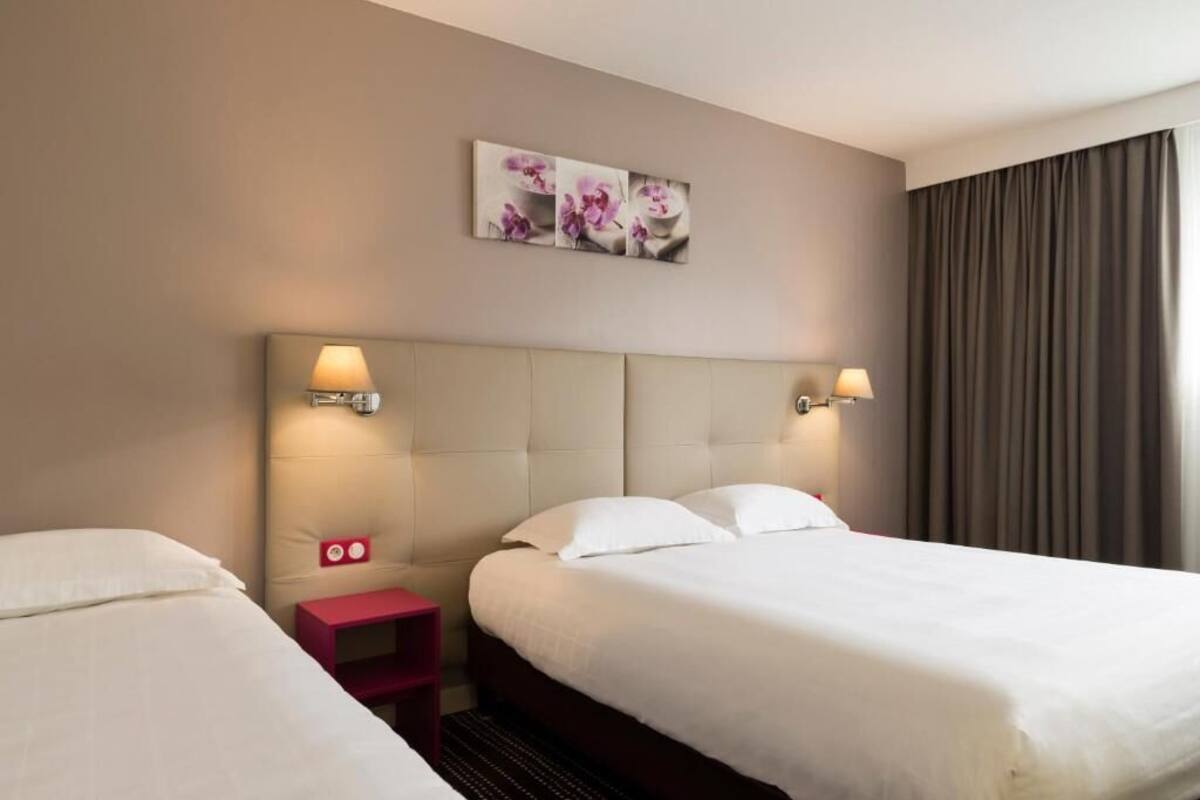 A hotel room with two beds, a headboard, wall-mounted lamps, a small red nightstand, and a floral artwork above the headboard.