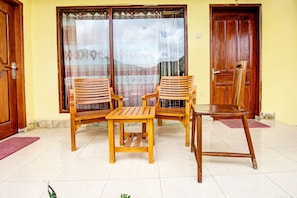 Um quarto com duas cadeiras de madeira, uma mesa pequena, uma porta de madeira e uma janela com cortinas.