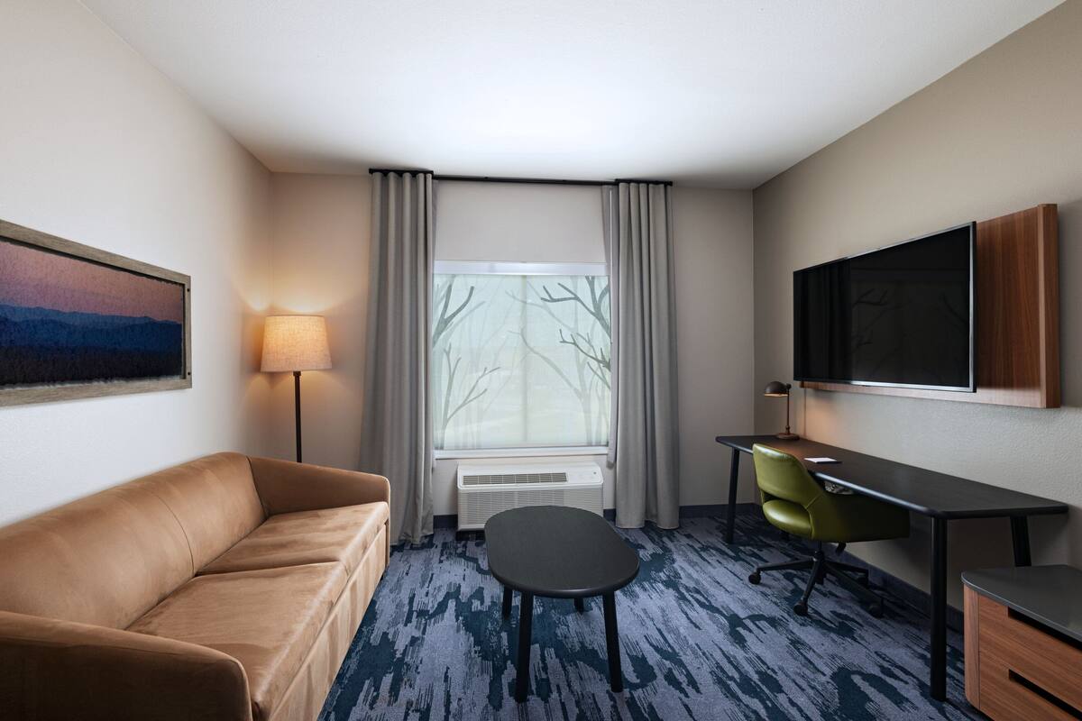 Une chambre d’hôtel équipée d’un canapé, d’un bureau, d’une chaise, d’un téléviseur à écran plat, d’une fenêtre avec des rideaux et d’un tableau au mur.