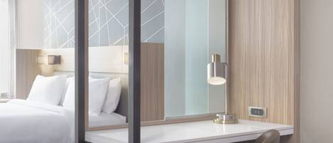 A hotel room with a bed, a desk, and a chair. There is a large mirror on the wall, a lamp on the desk, and a window with curtains.