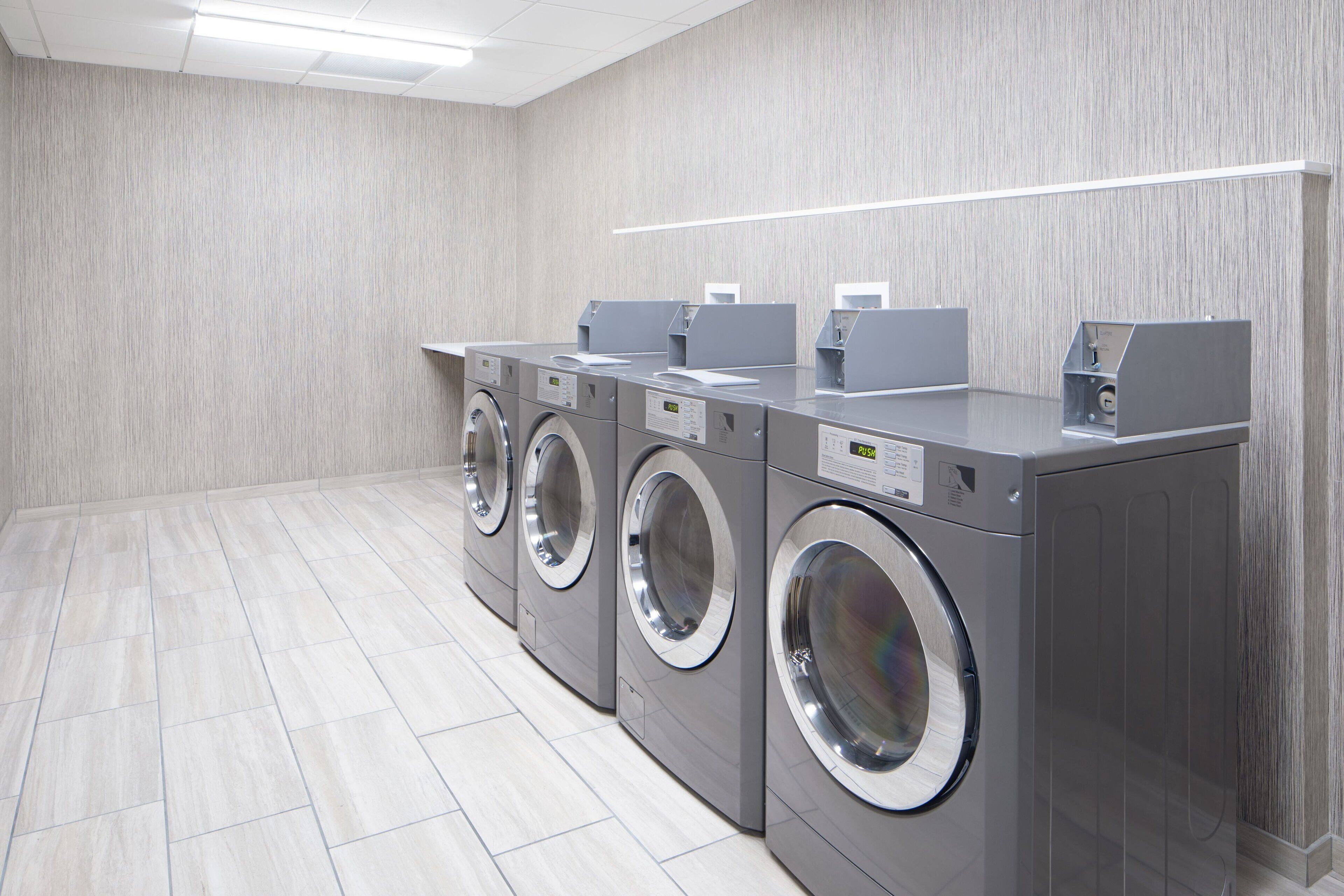 A row of grey, stainless steel front-loading washing machines in a modern, tiled laundry room with a light wood-textured wall and a linear light fixture above.
