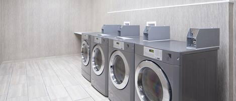 A row of grey, stainless steel front-loading washing machines in a modern, tiled laundry room with a light wood-textured wall and a linear light fixture above.