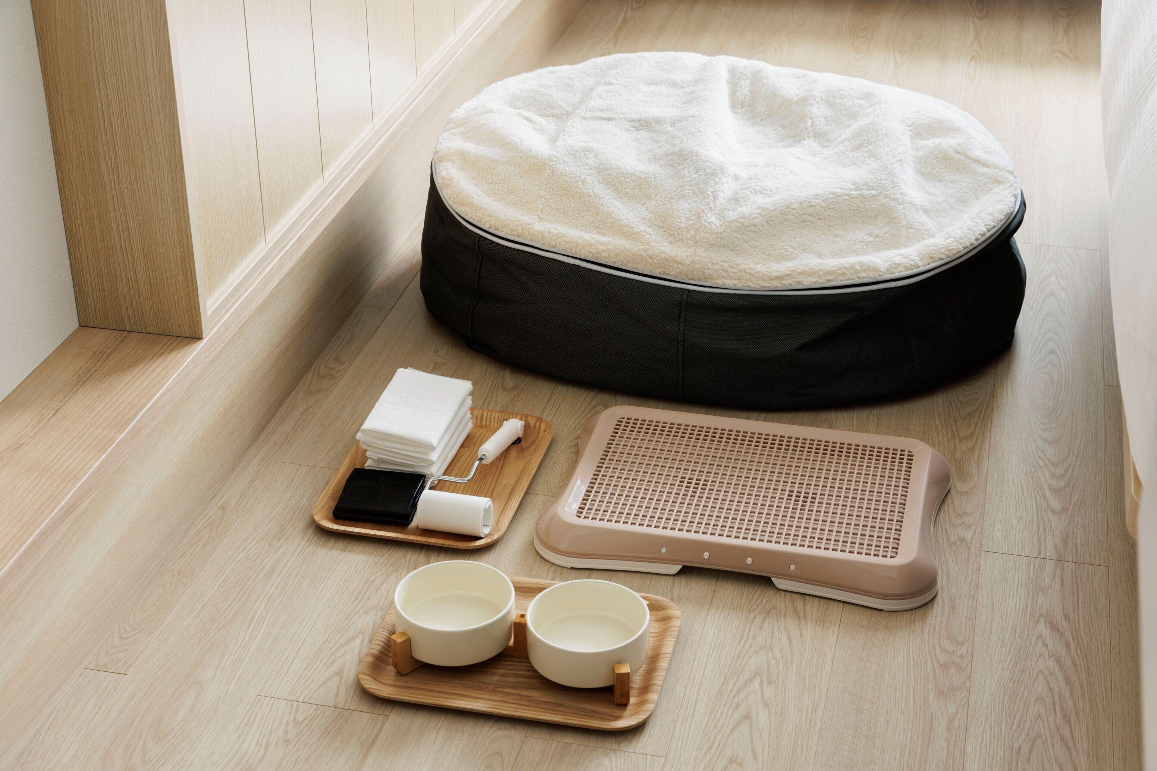 Una habitación con una cama redonda negra para mascotas, una cama beige para mascotas, una bandeja de madera con dos cuencos y un cepillo, y una alfombra beige para mascotas.