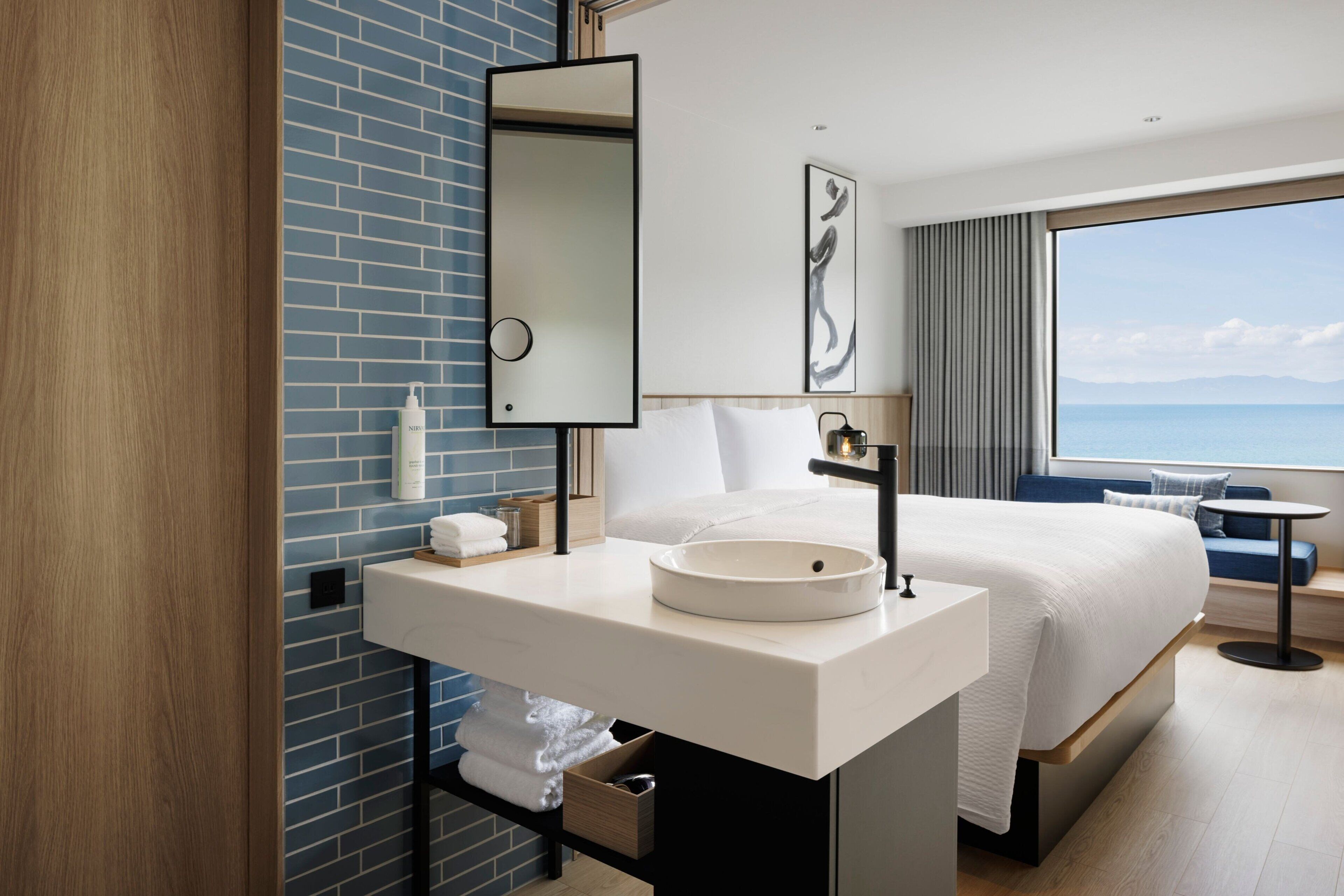 A modern hotel room with a large bed, a white bathroom with a round sink, and a view of the sea through a large window.