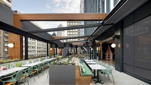 An outdoor dining area with long tables and chairs, surrounded by plants and a wooden bench.