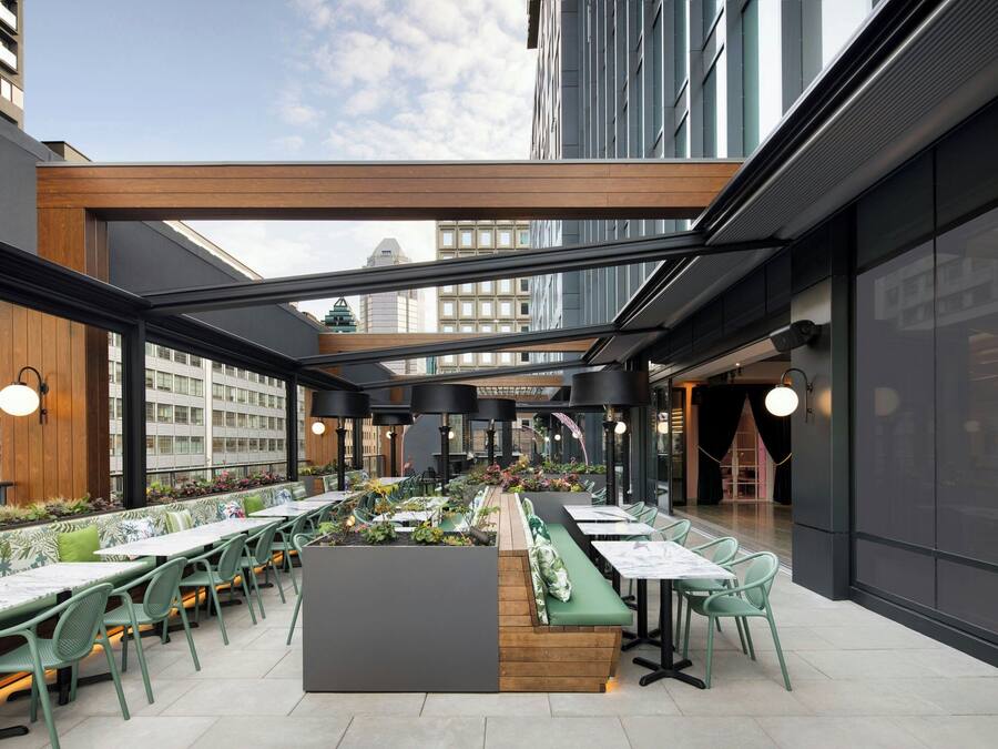 An outdoor dining area with long tables and chairs, surrounded by plants and a wooden bench.