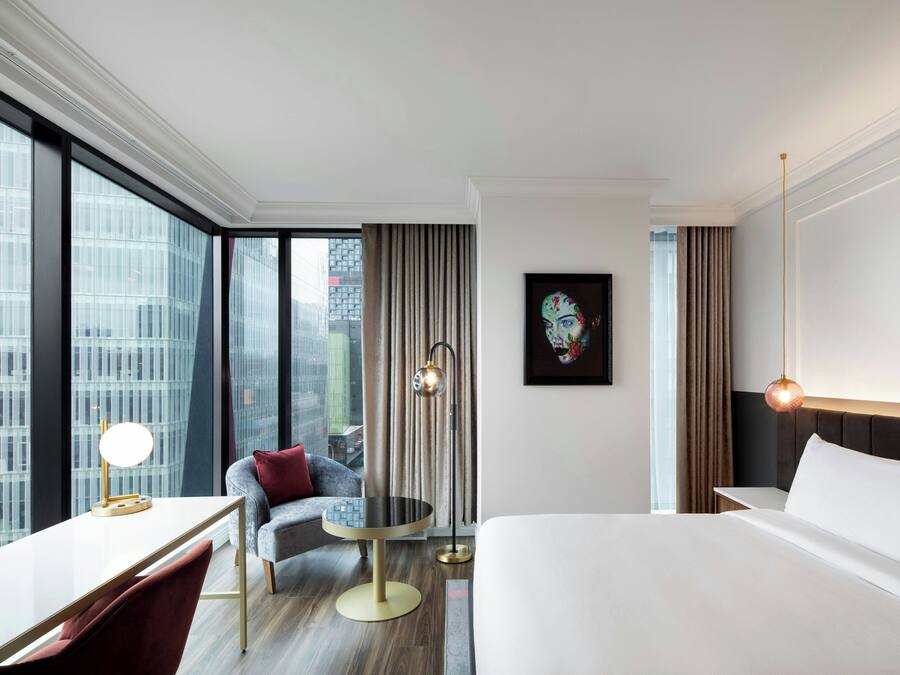 Habitación de hotel moderna con una cama grande, un escritorio con lámpara, una mesa redonda y vistas a la ciudad a través de amplios ventanales.