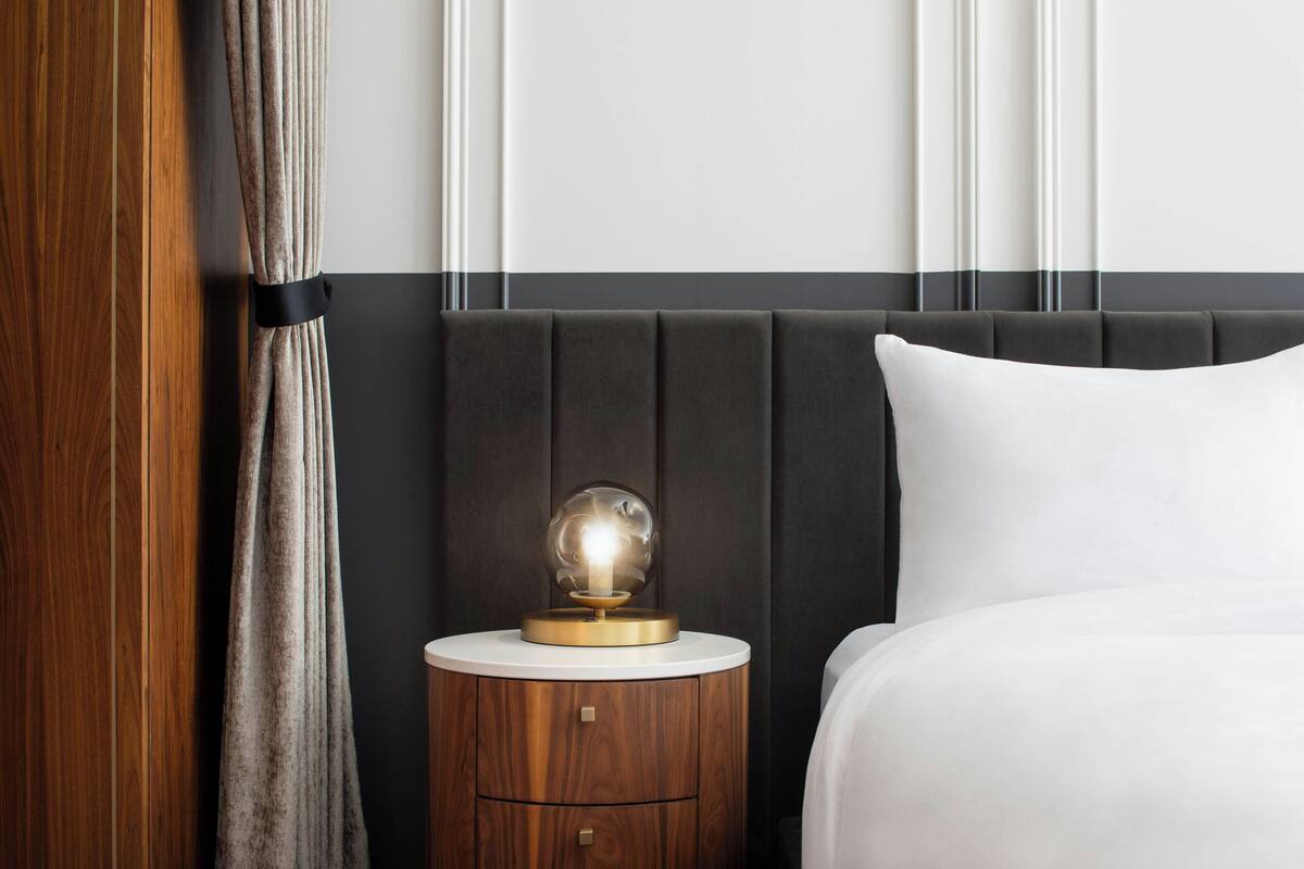 Una cama bien hecha con una almohada blanca, una mesita de noche redonda con una lámpara y una mesita auxiliar de madera.