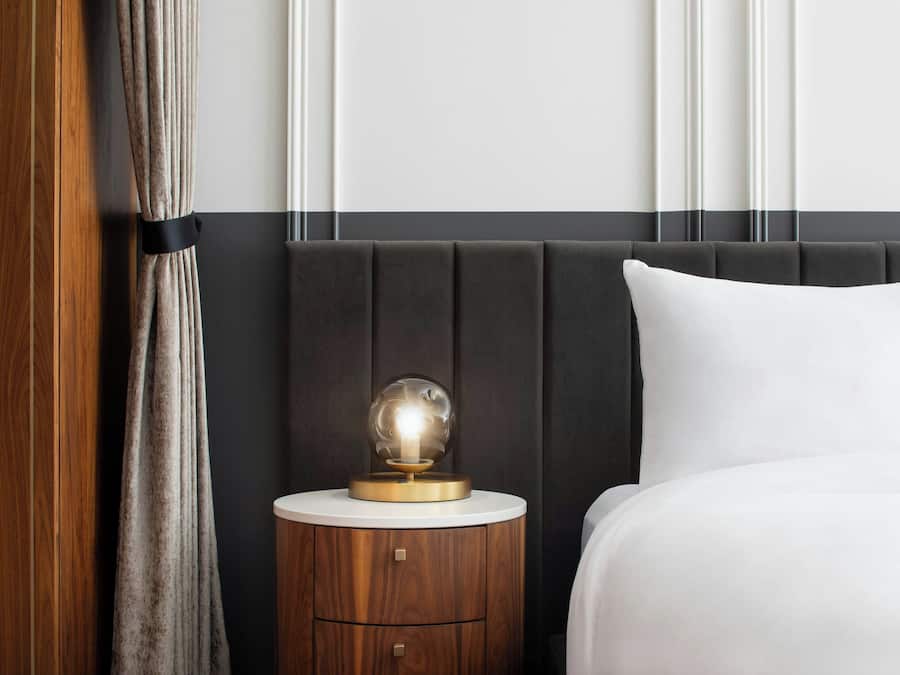 Una cama bien hecha con una almohada blanca, una mesita de noche redonda con una lámpara y una mesita auxiliar de madera.