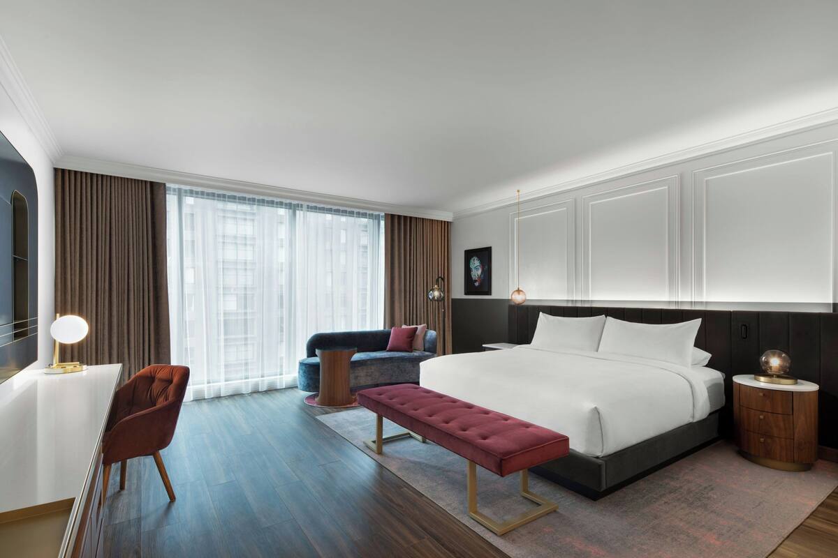 Una habitación de hotel moderna con una cama grande, un escritorio, una silla, un banco y un sofá.
