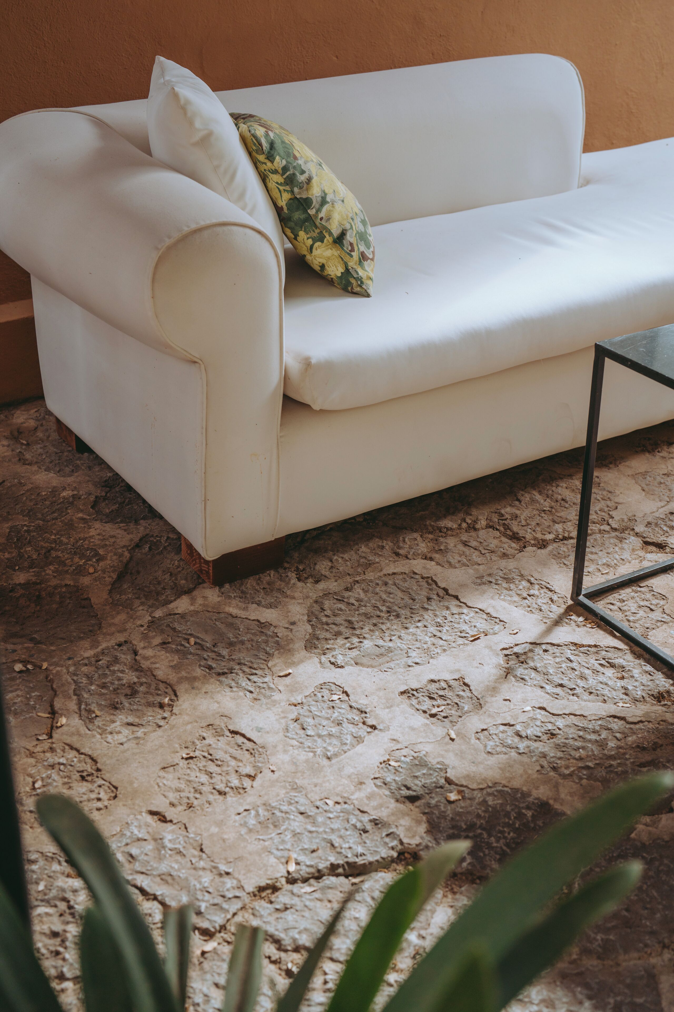A white sofa with a floral pillow against a textured stone floor, with a metal frame visible in the foreground.