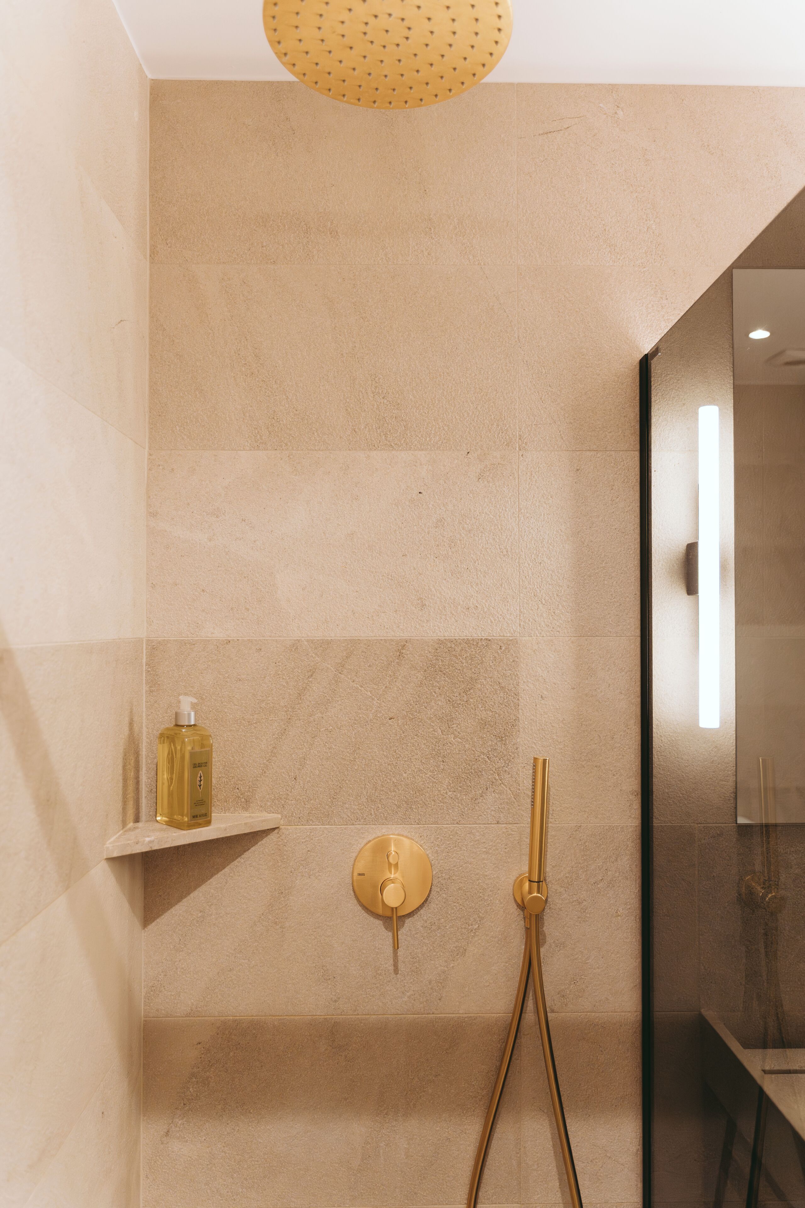 A modern bathroom with a shower, a gold-colored faucet, and a bottle of shampoo on a shelf.