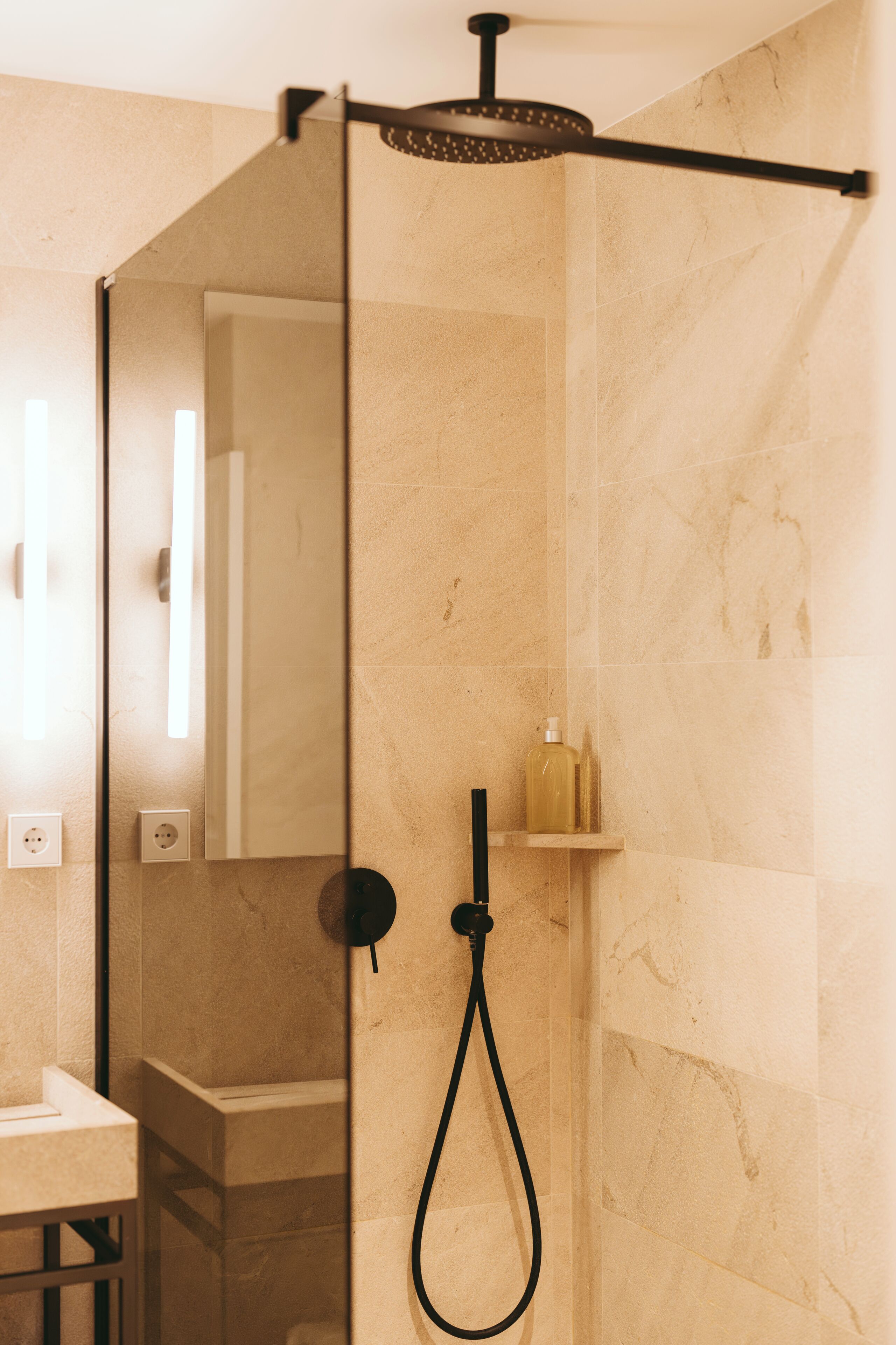 A modern bathroom with a glass-enclosed shower, marble walls, and a small shelf with toiletries.