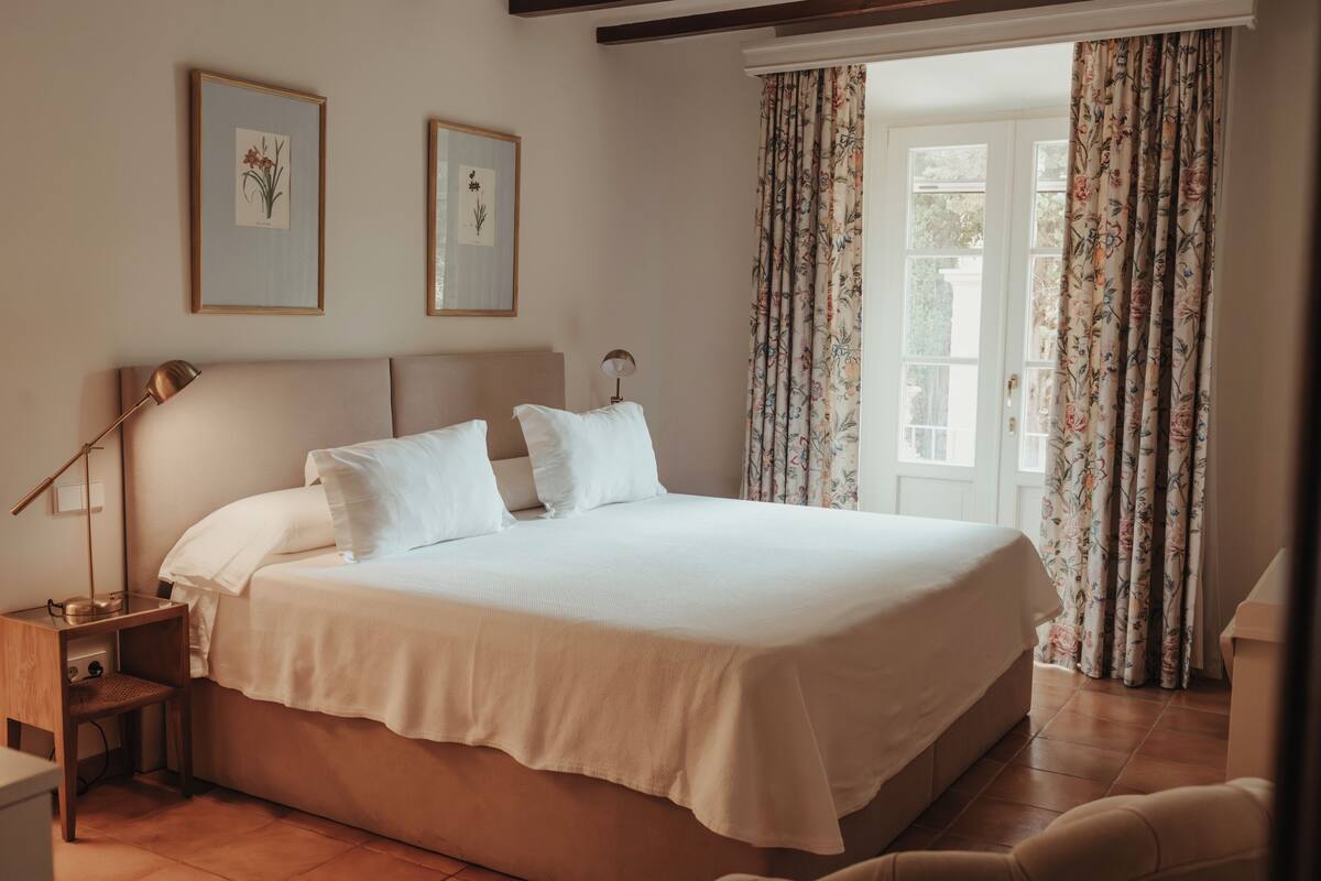 A neatly made bed with white linens, a bedside lamp, and framed botanical prints on the wall.