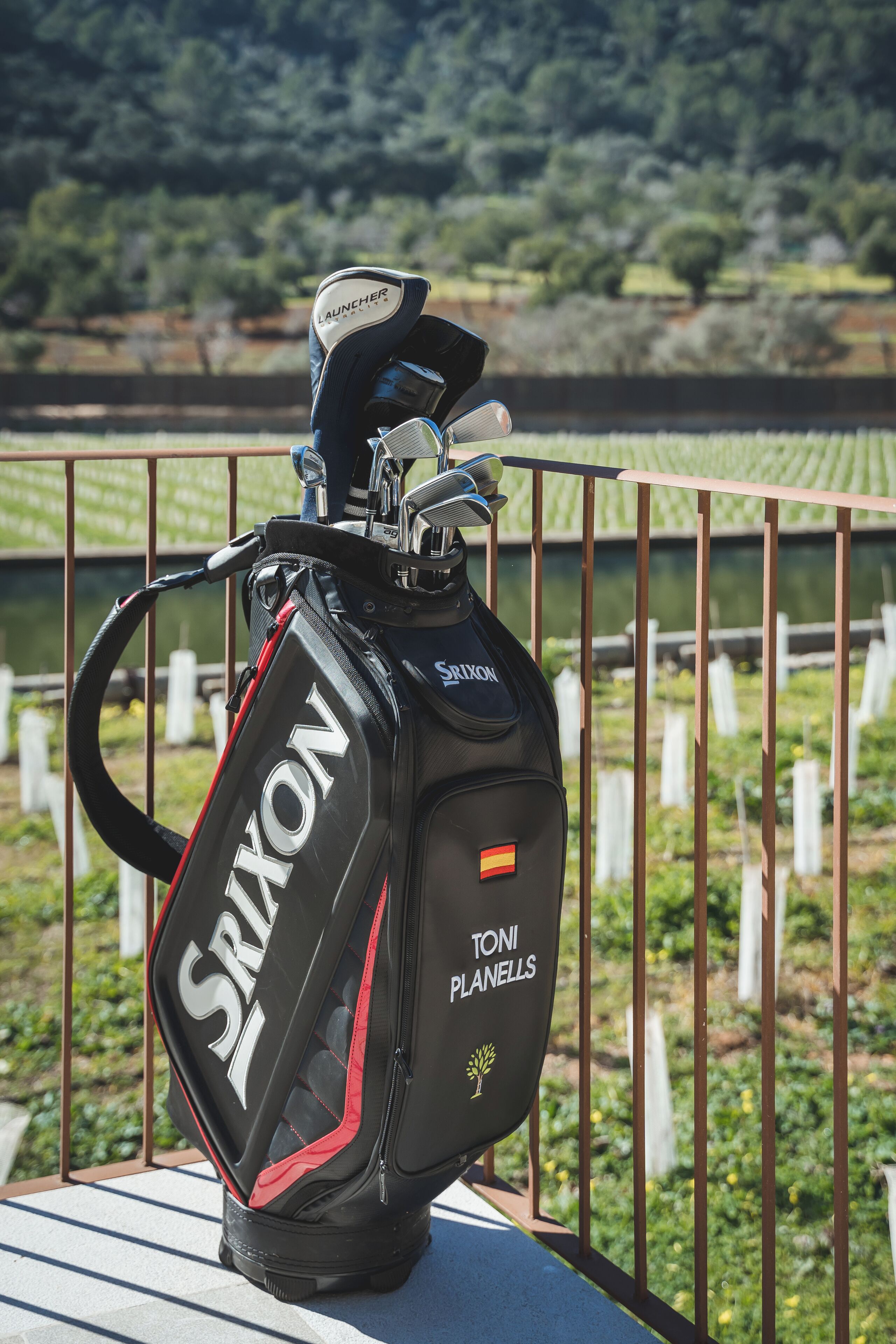 A golf bag with the brand name "Srixon" and the text "TONI PLANELLS" on it, placed on a railing with a view of a landscaped area and a body of water in the background.