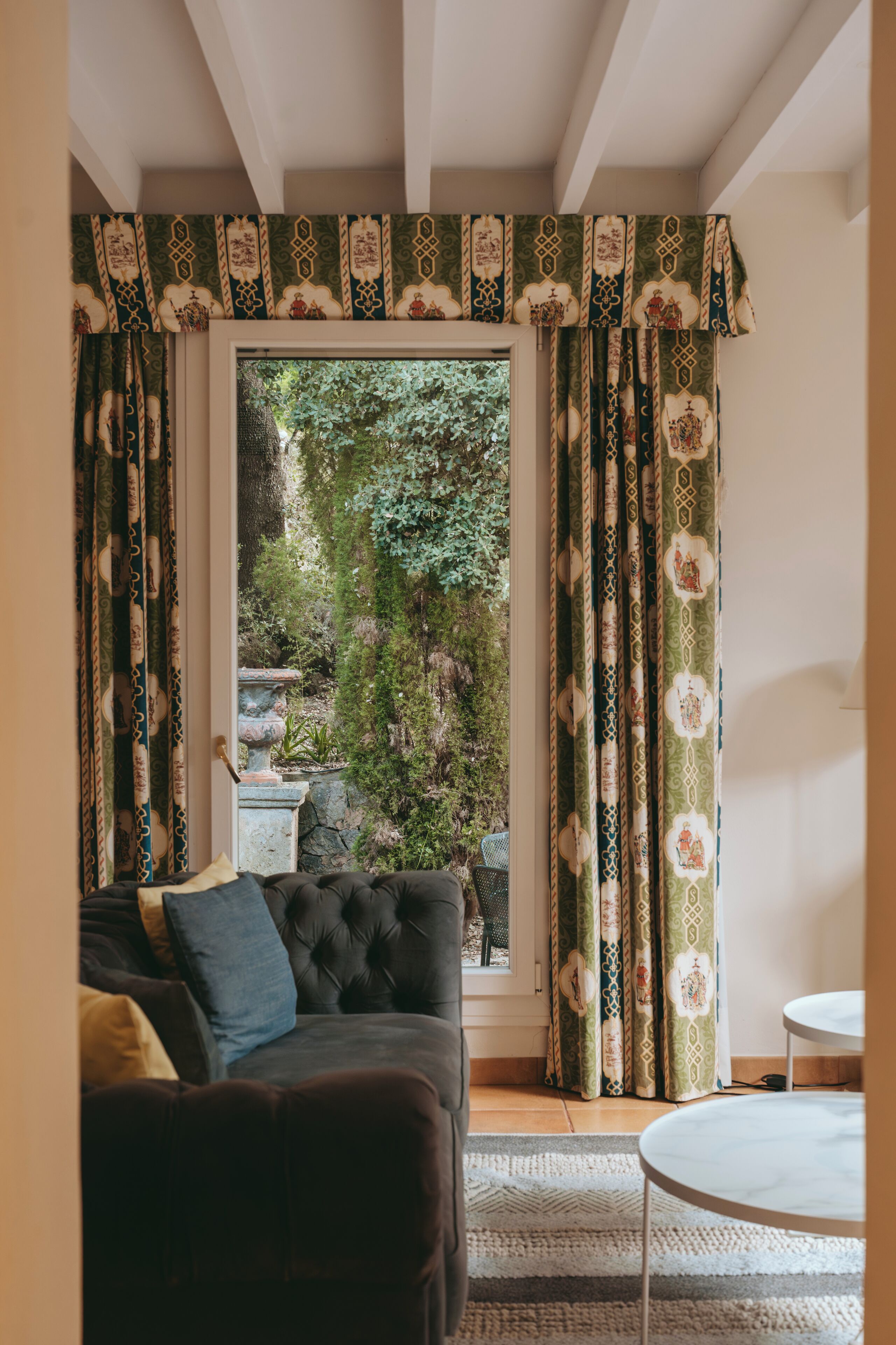 A cozy living room with a dark sofa, a small round table, and a view of a garden through a window with patterned curtains.