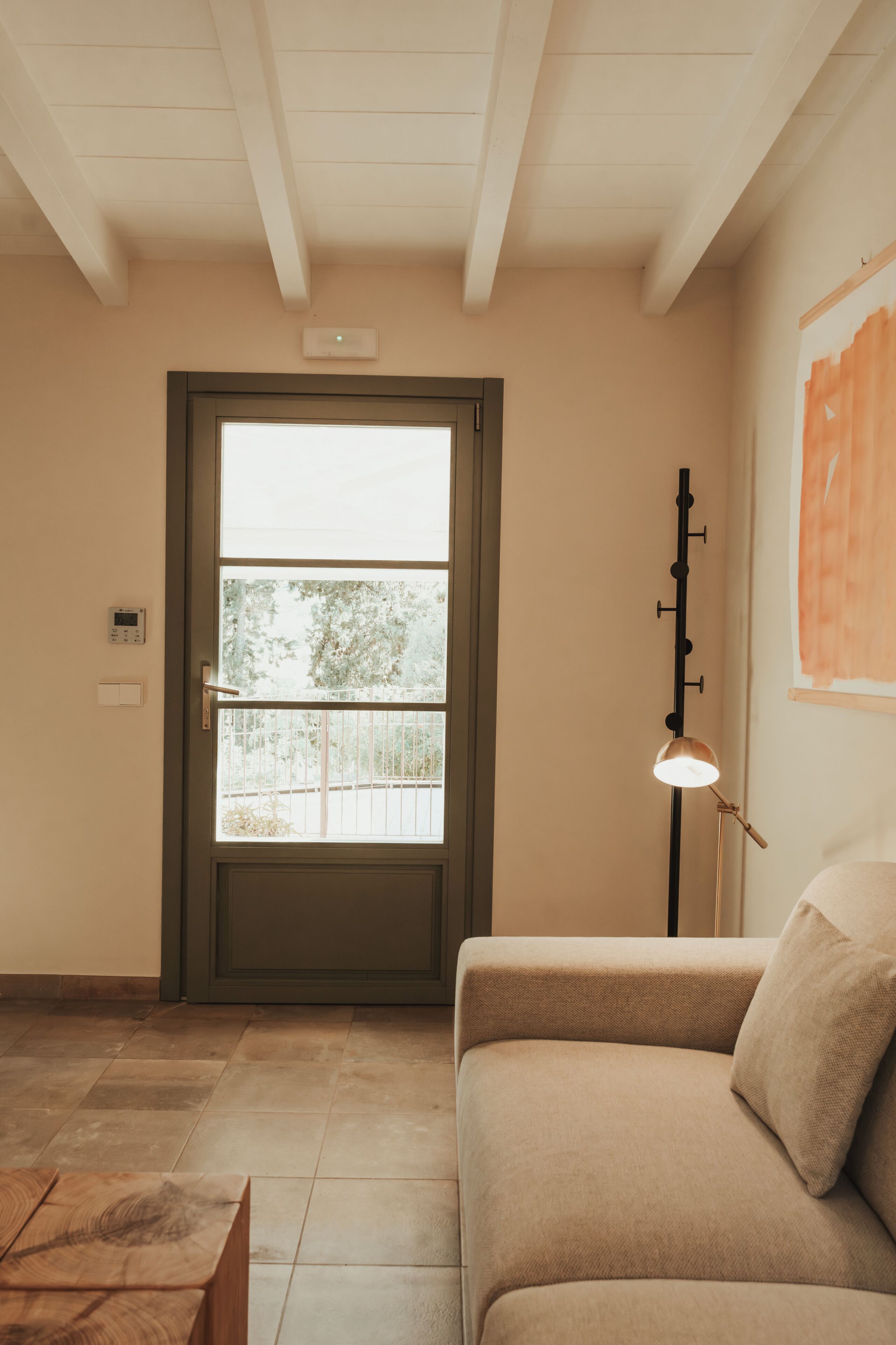 A room with a beige sofa, a door with a view to the outside, a floor lamp, and a thermostat on the wall.