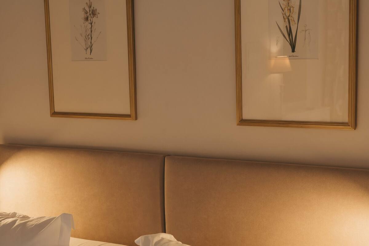 A neatly made bed with white linens and pillows, a headboard, and two framed botanical prints on the wall.