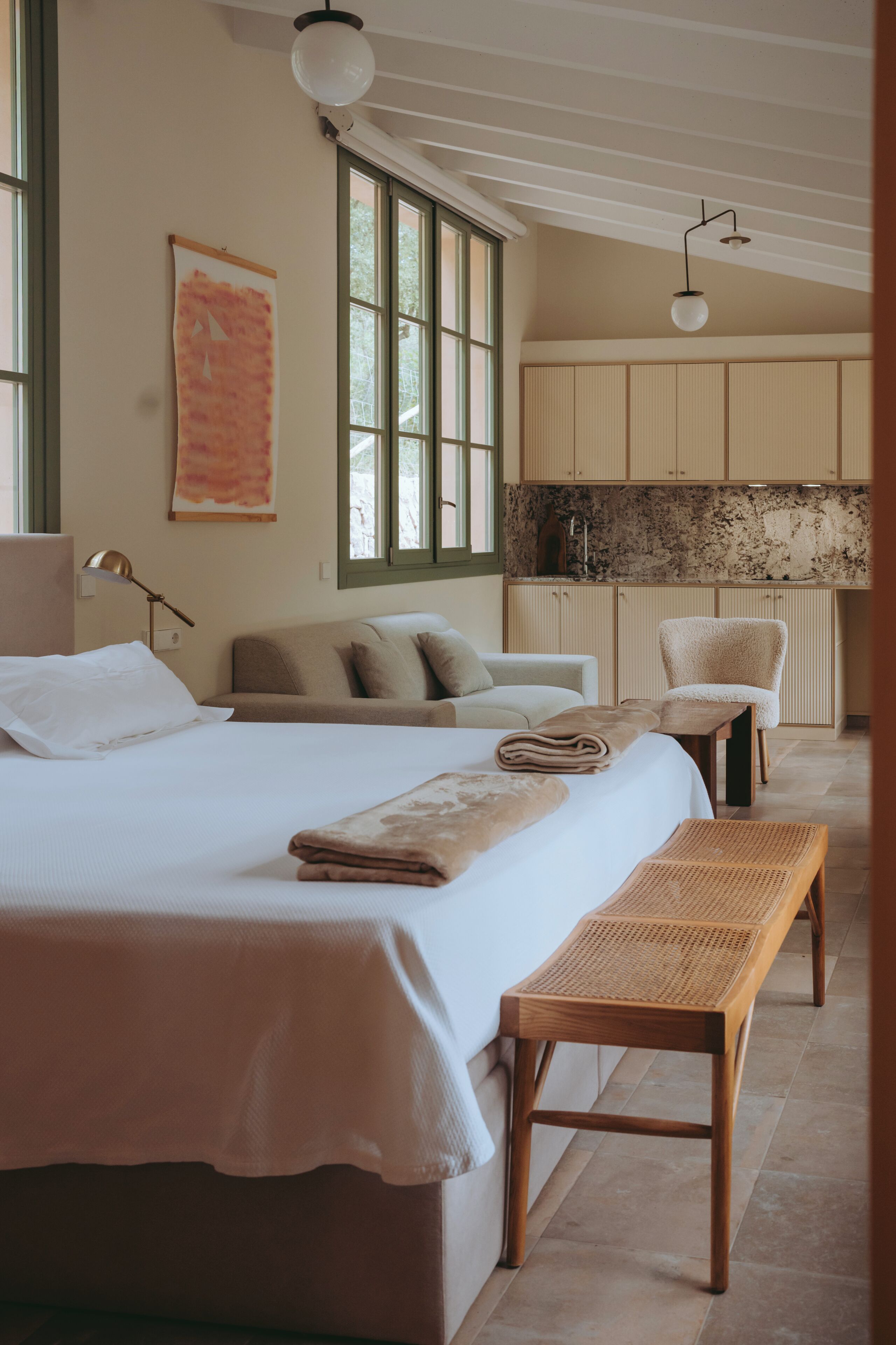 Un dormitorio con una cama grande, un banco, una silla y una kitchenette al fondo.
